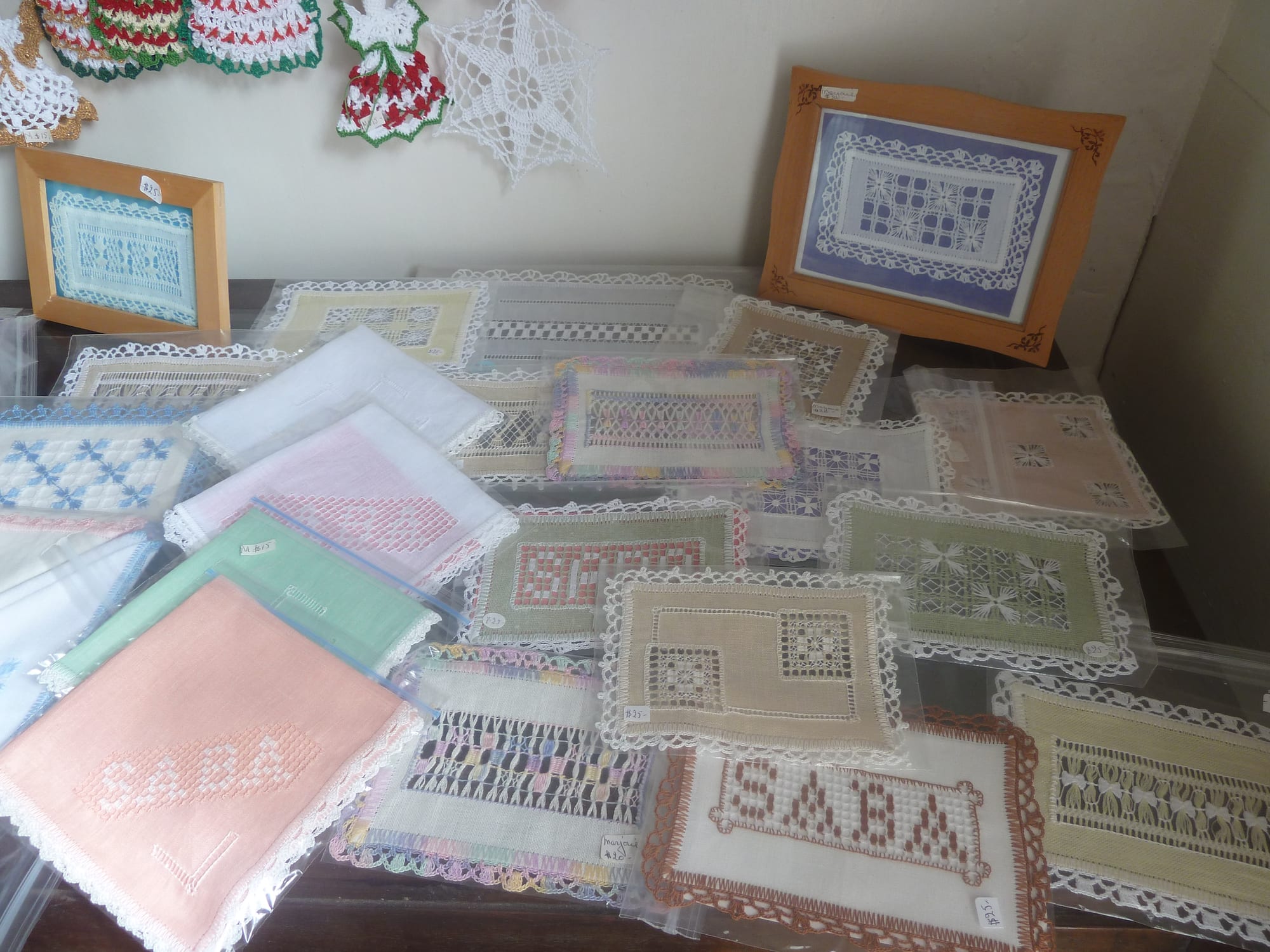 A table covered with squares of embroidered drawn-thread lace in white, pink, green, blue and brown, some in plastic sleeves, two with the word SABA worked into the pattern, and framed pieces behind