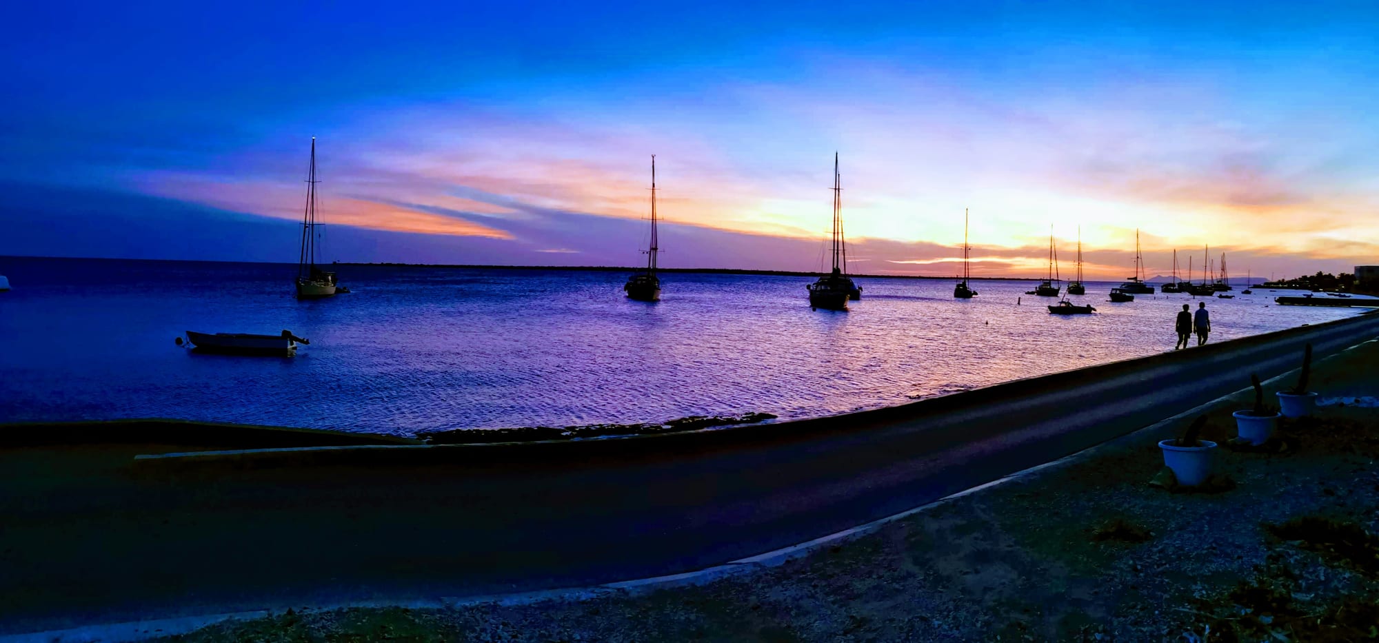 Sailboats moored in the calm water off Kralendijk, Bonaire, at sunset, with two people walking along the boulevard