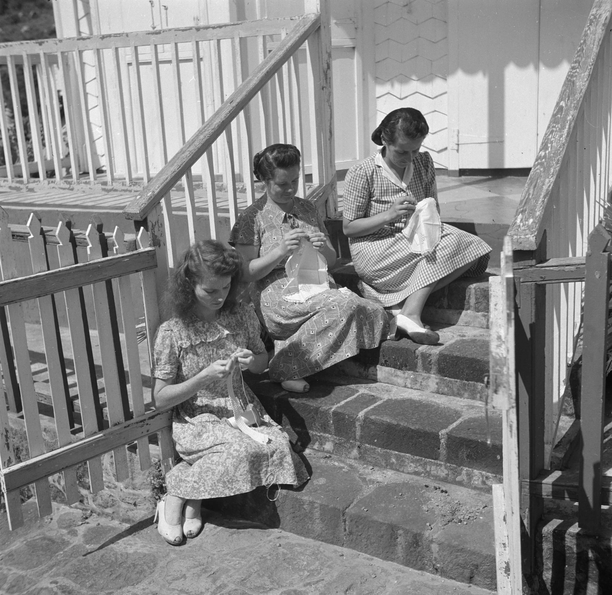 Black-and-white photograph of three women in patterned dresses sitting on stone steps beside a white wooden house, each bent over a piece of white needlework