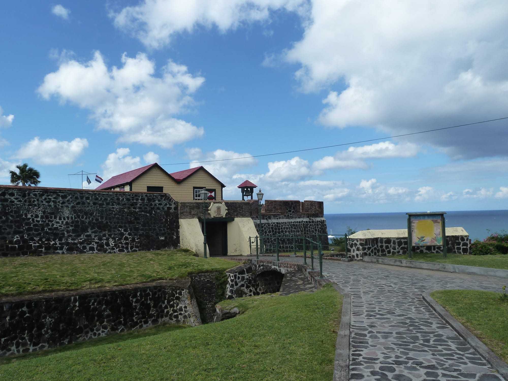 A restored stone fort with cannon and a Dutch flag on a cliff-top above a blue bay
