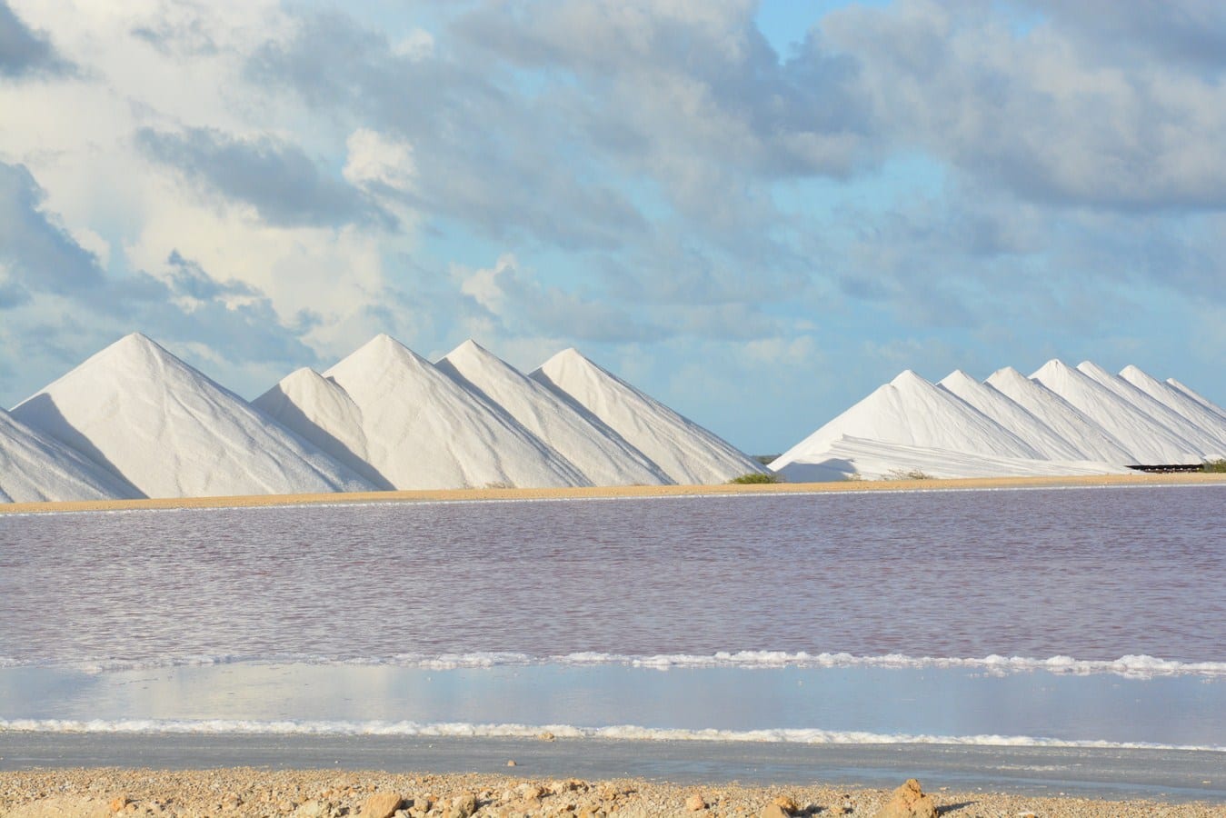 White pyramids of harvested sea salt beside a shallow pink-tinted lagoon under a wide sky