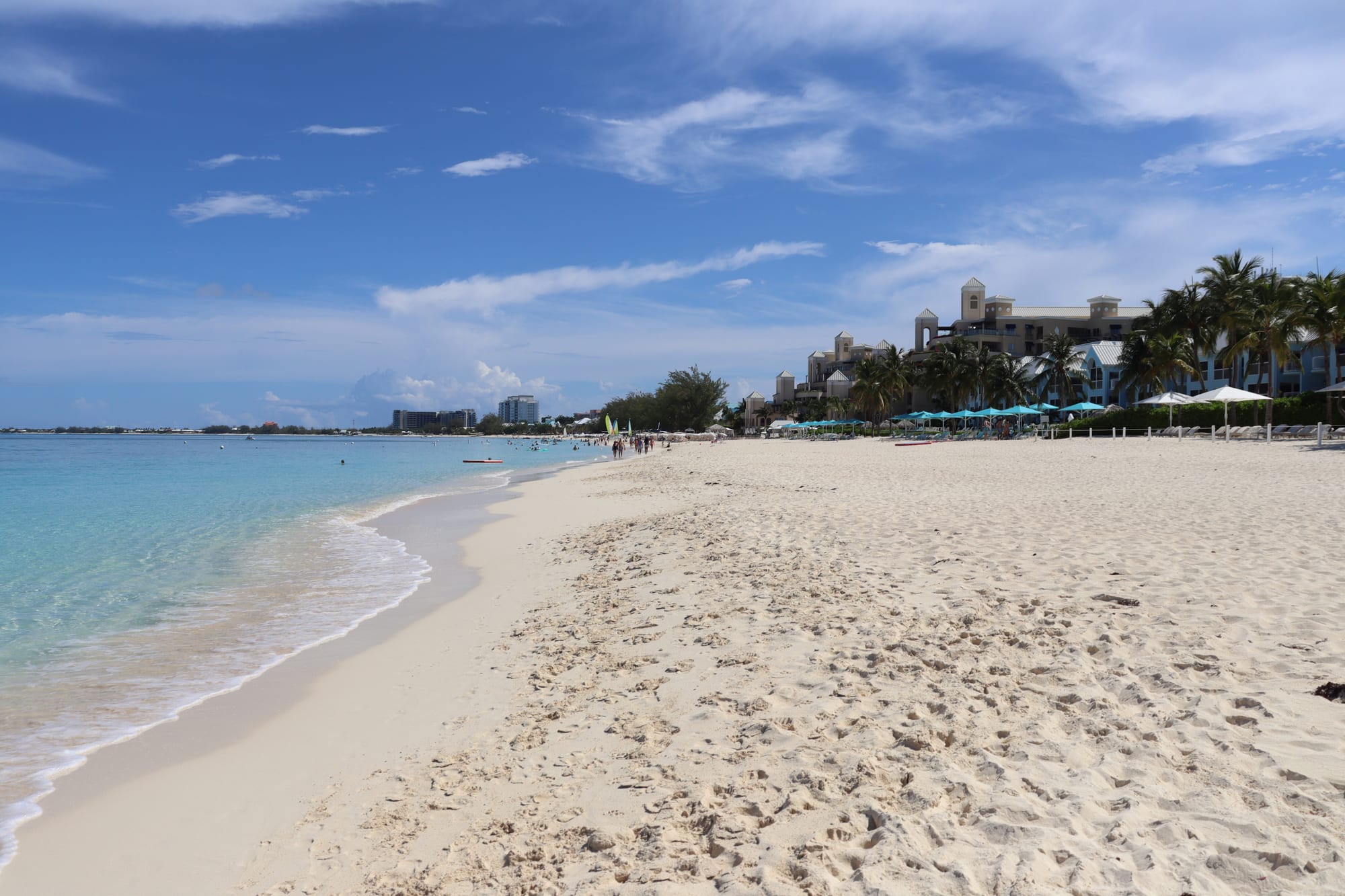 A long straight beach of pale sand and calm turquoise water, resorts set back among palms