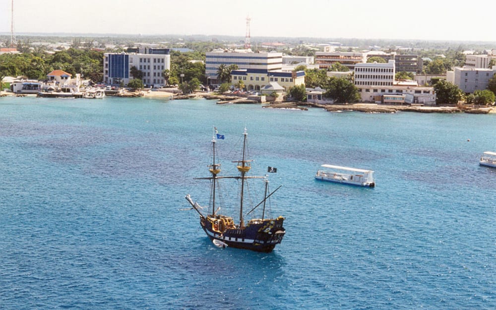A low-rise waterfront town of office buildings and shops beside a calm harbour with a cruise ship offshore