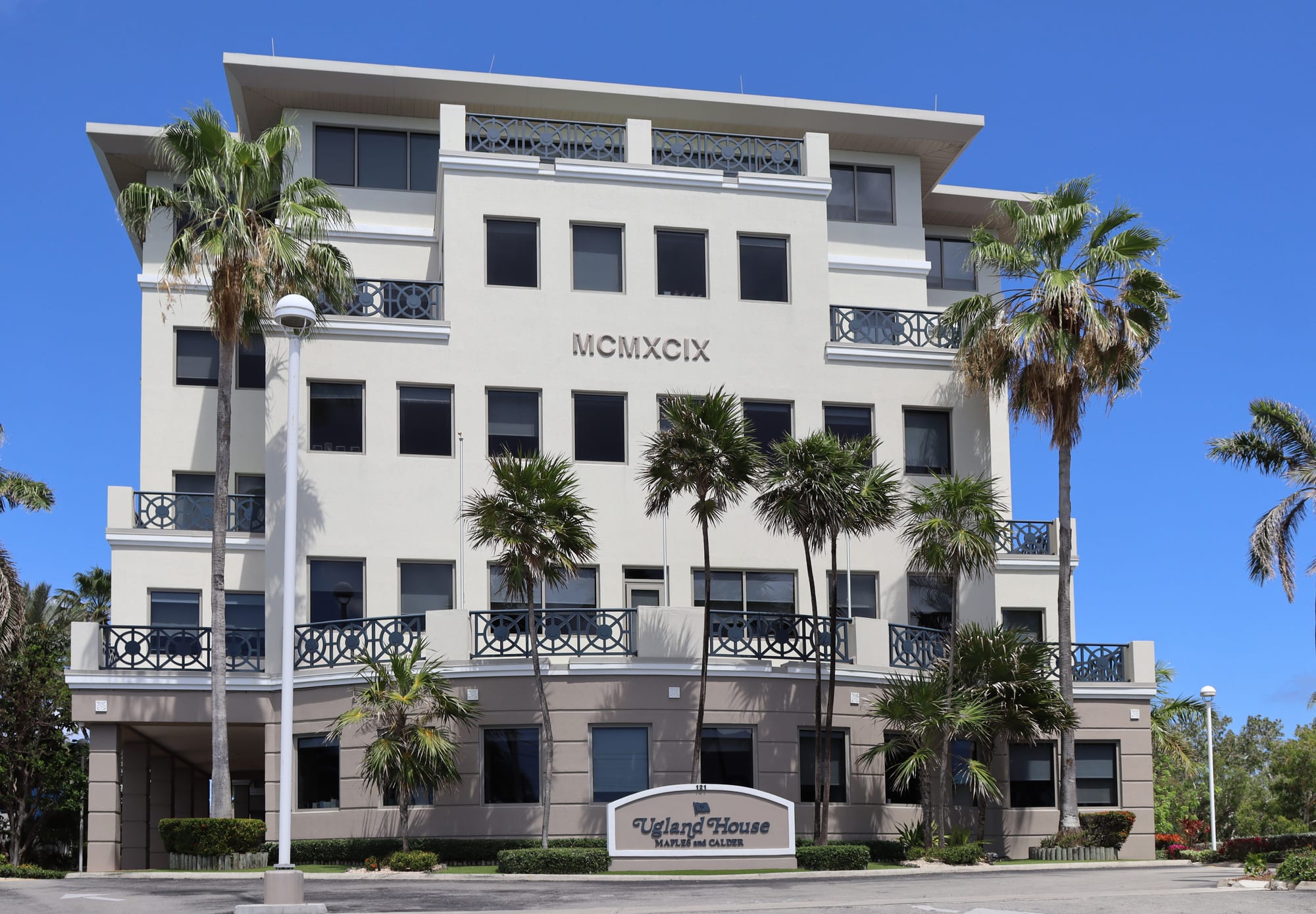 A five-storey cream office building with dark iron balconies and the Roman numerals MCMXCIX on its front, framed by palm trees, with a sign reading Ugland House at the entrance