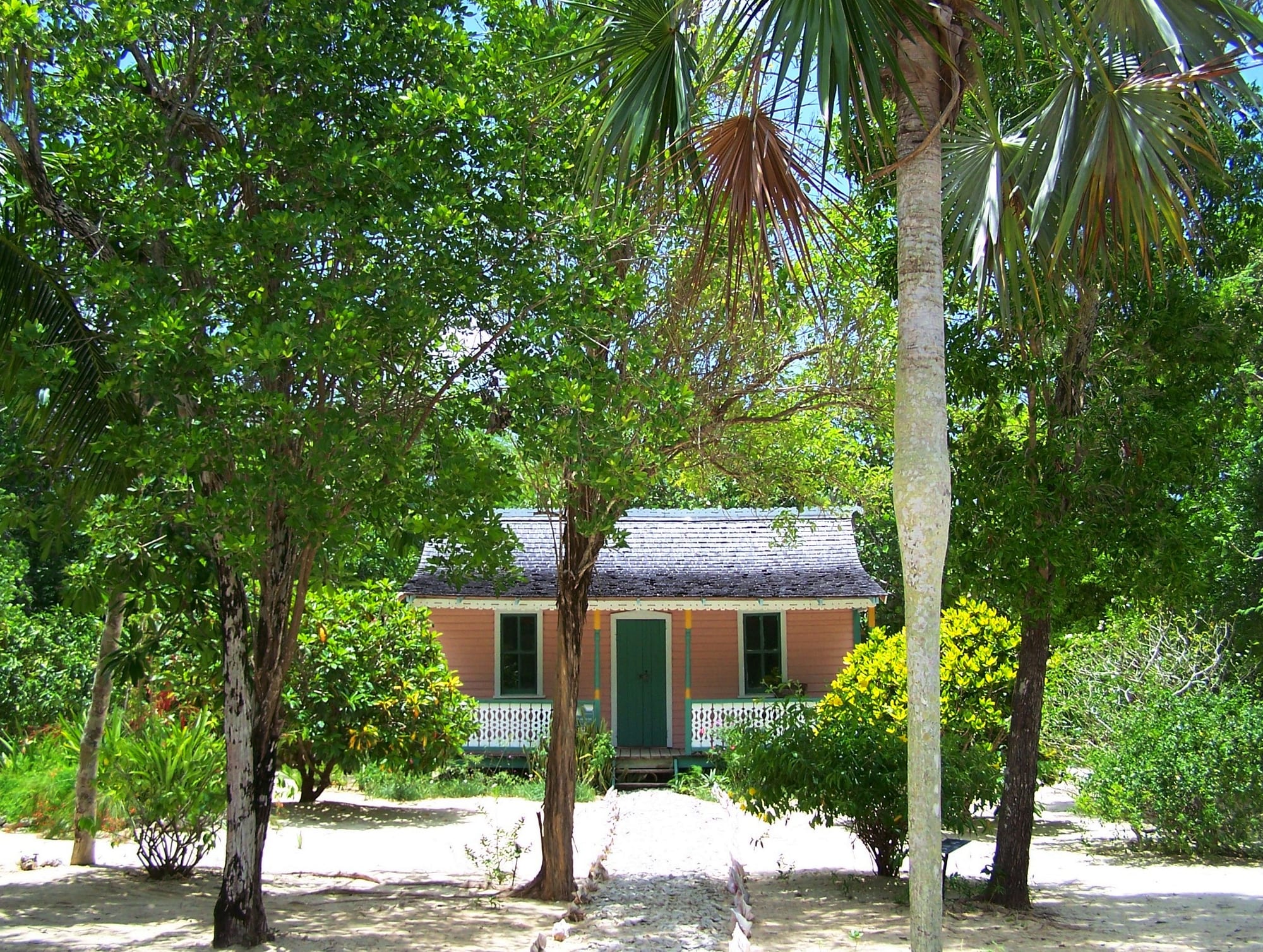 A small salmon-pink wooden cottage with a grey shingled roof, a green front door and white porch railings, set back in a yard of white sand among trees, with a tall pale-trunked fan palm in the foreground