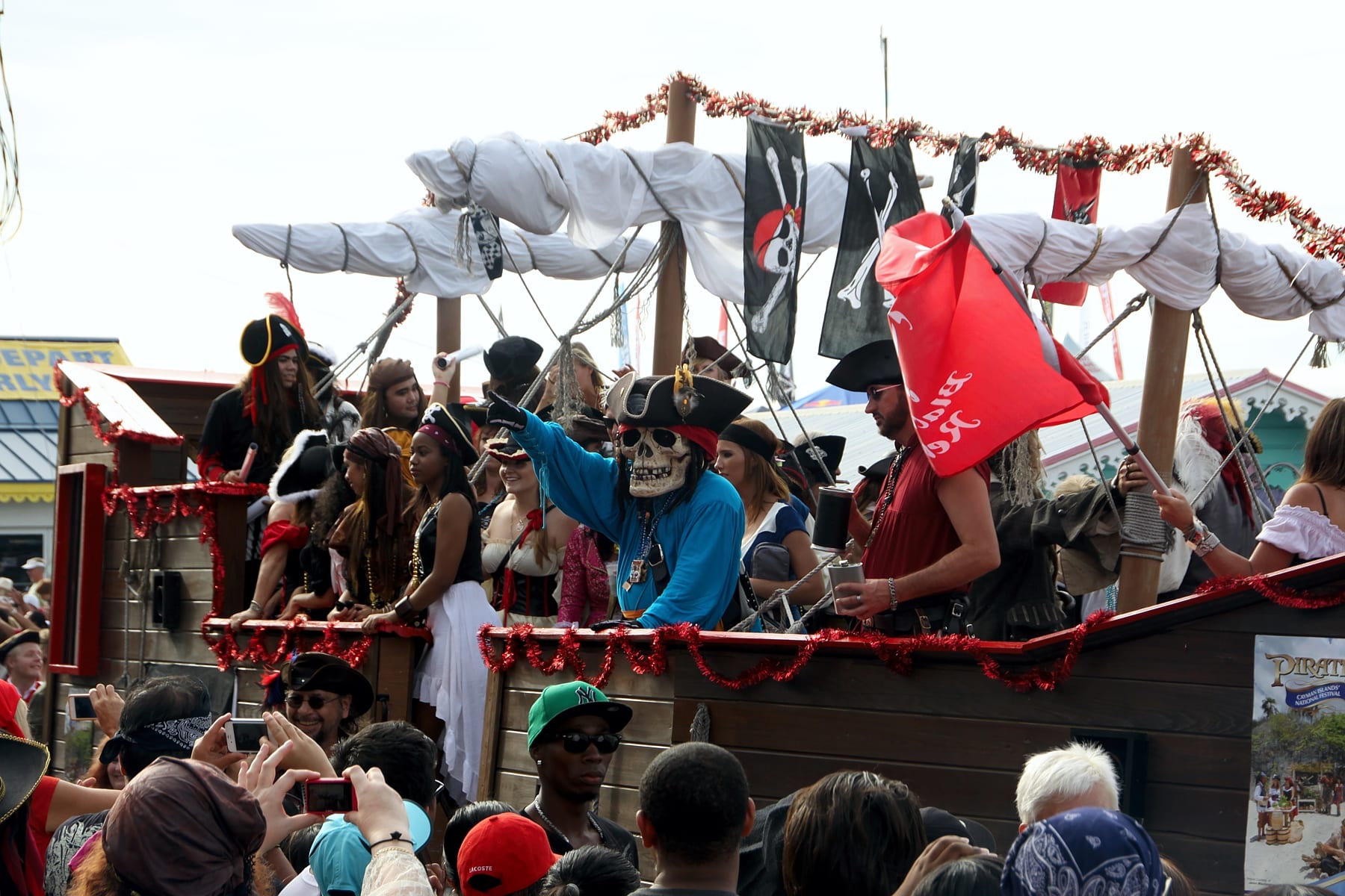 A float built as a pirate ship, with white sails, black skull-and-crossbones flags and red tinsel, carries costumed pirates past a crowd of spectators taking photographs