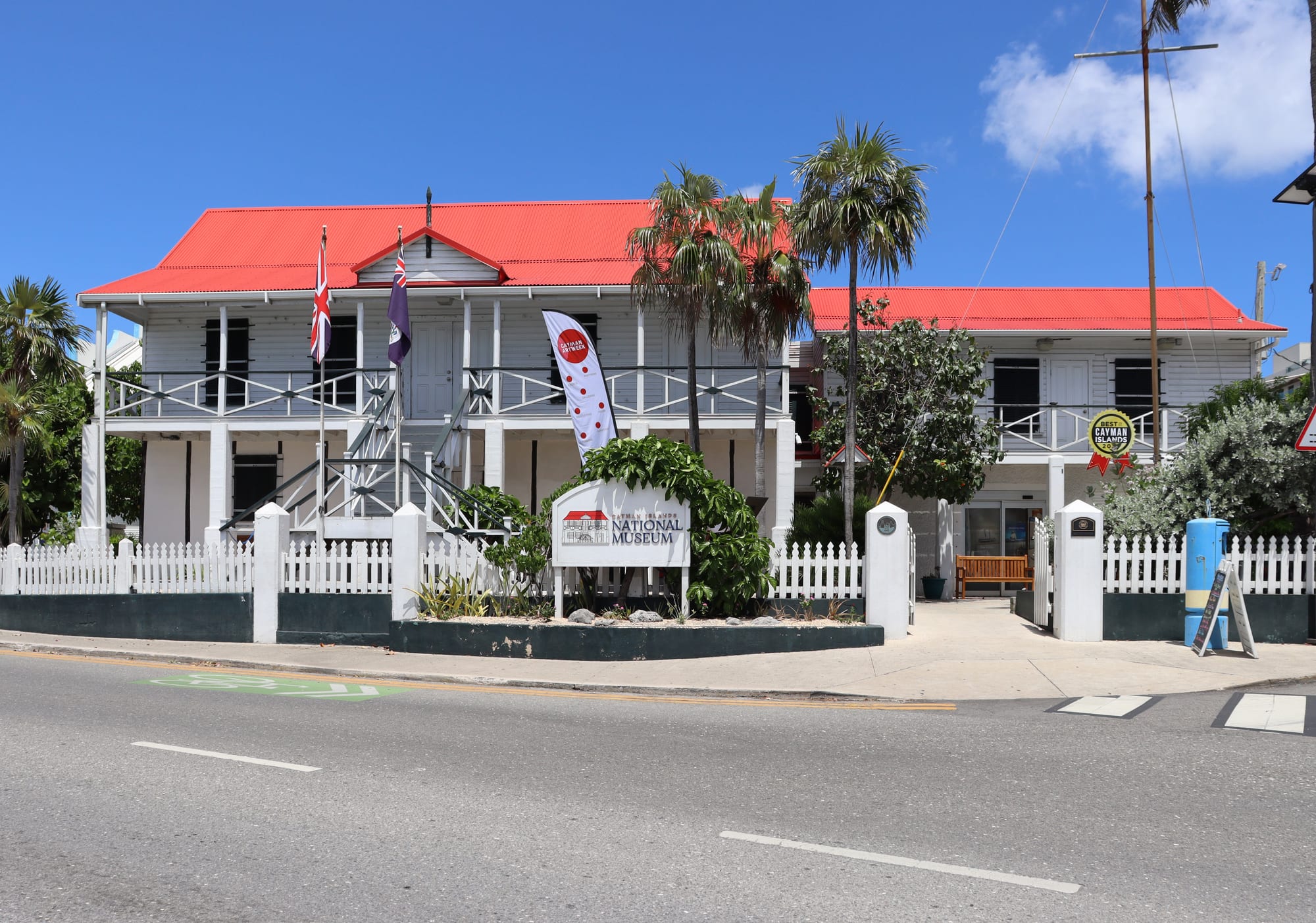 A two-storey white clapboard building with a bright red corrugated roof, wooden verandas and a white picket fence, with a National Museum sign and flags in front under a blue sky