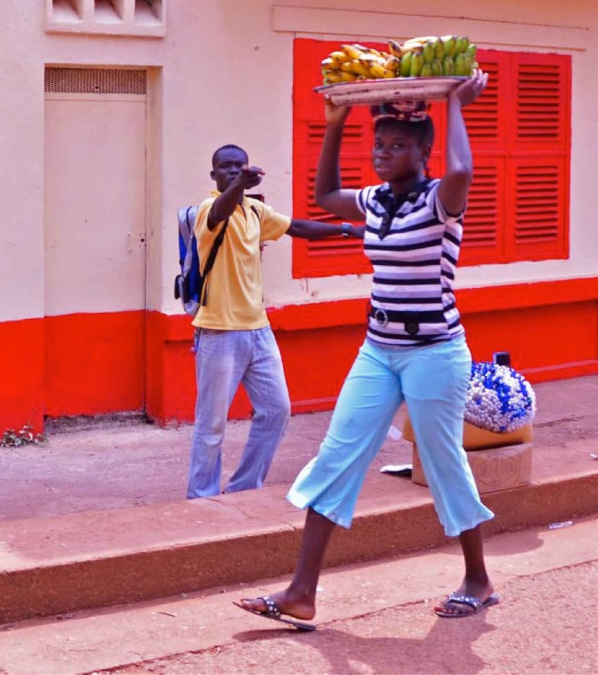A young woman carrying a tray of bananas on her head while walking down a street, with a man in the background