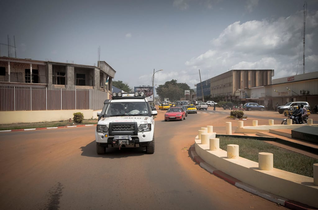 A roundabout on a Bangui street with a four-wheel-drive, cars, a motorbike and pedestrians under a bright sky