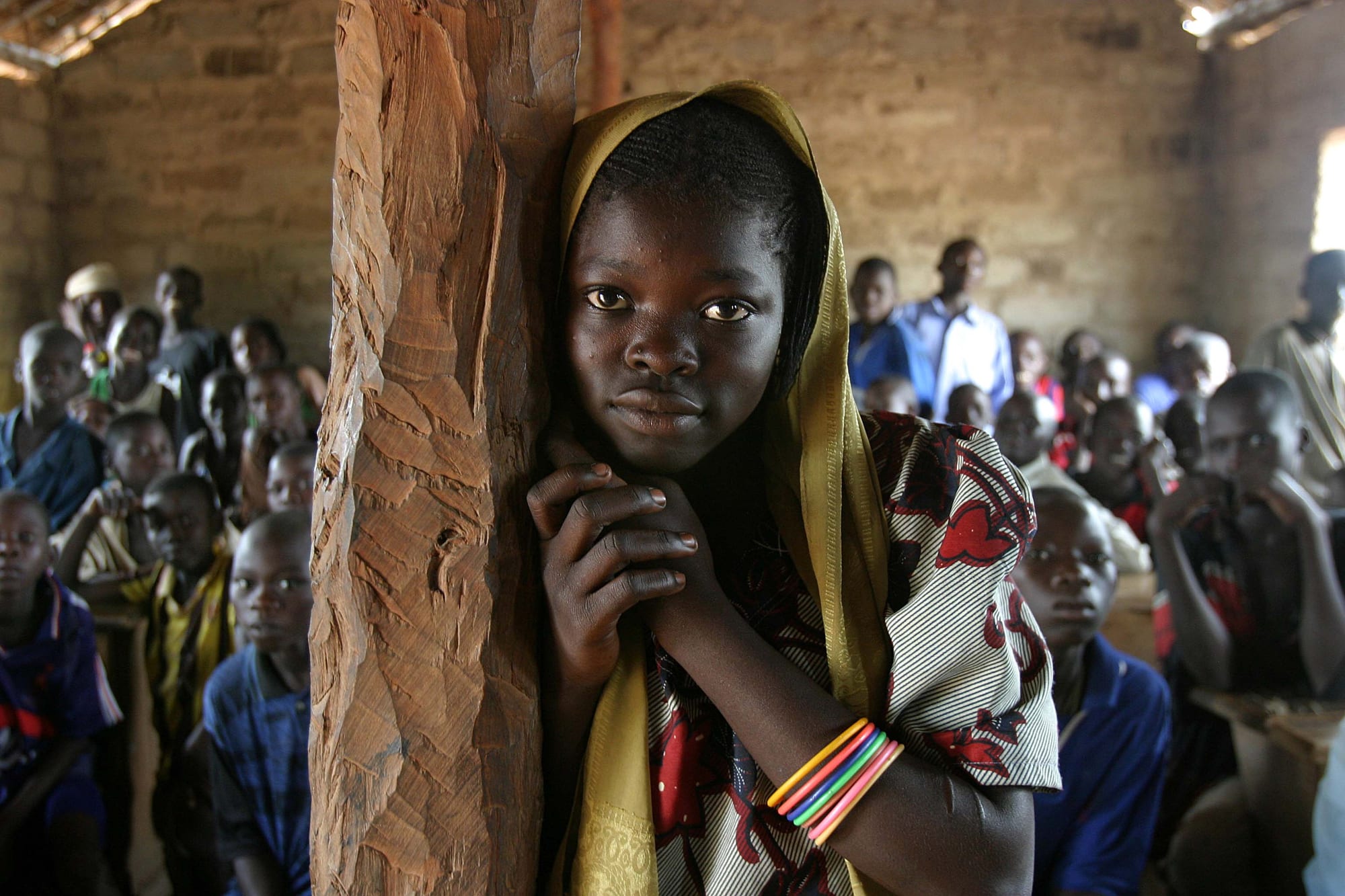 A crowded classroom of Central African schoolchildren, with a girl standing by a wooden support beam in the foreground