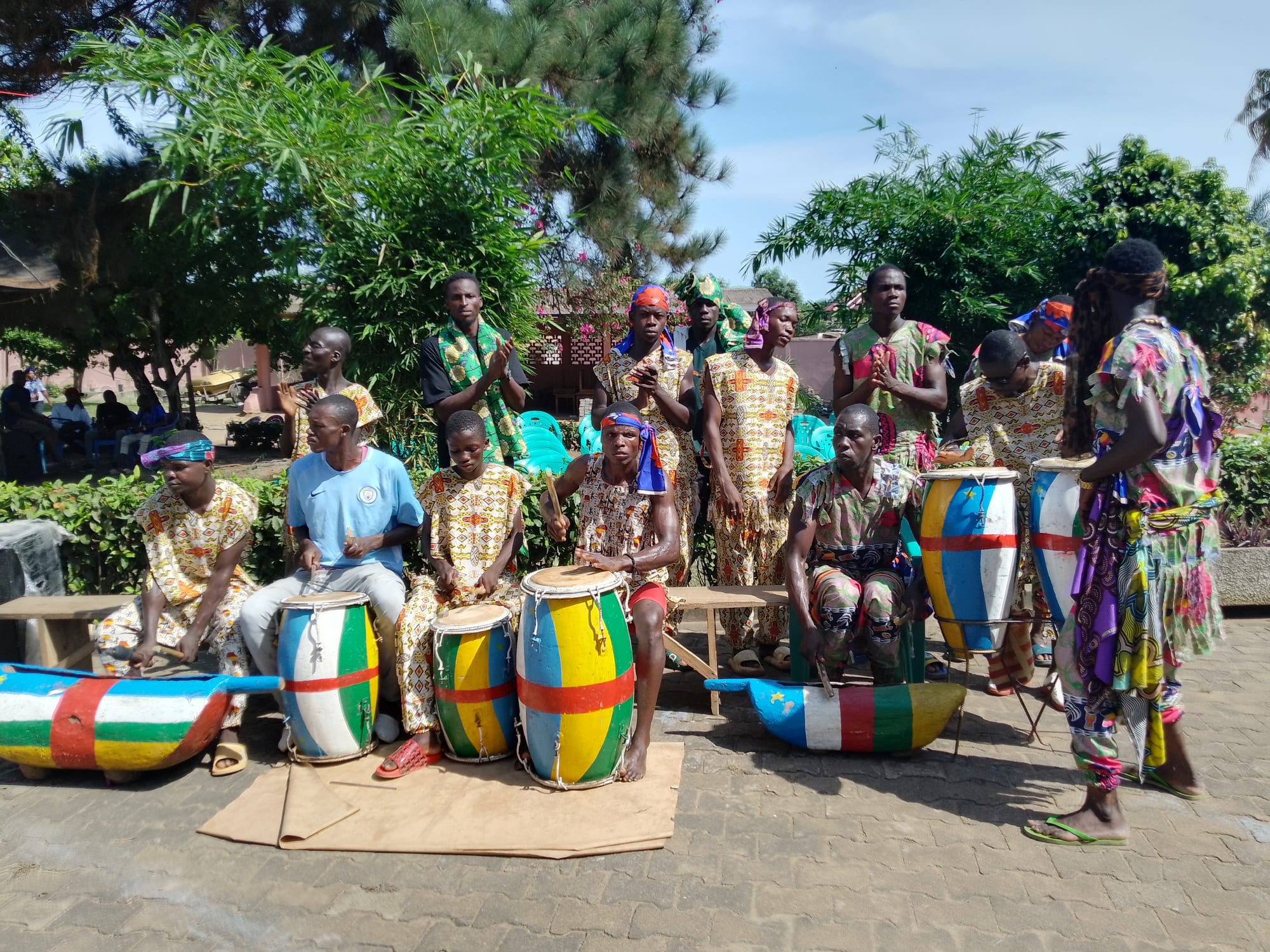 A traditional music and dance group in matching patterned clothes and headbands, seated and standing behind tall drums and slit drums painted in blue, white, green, yellow and red