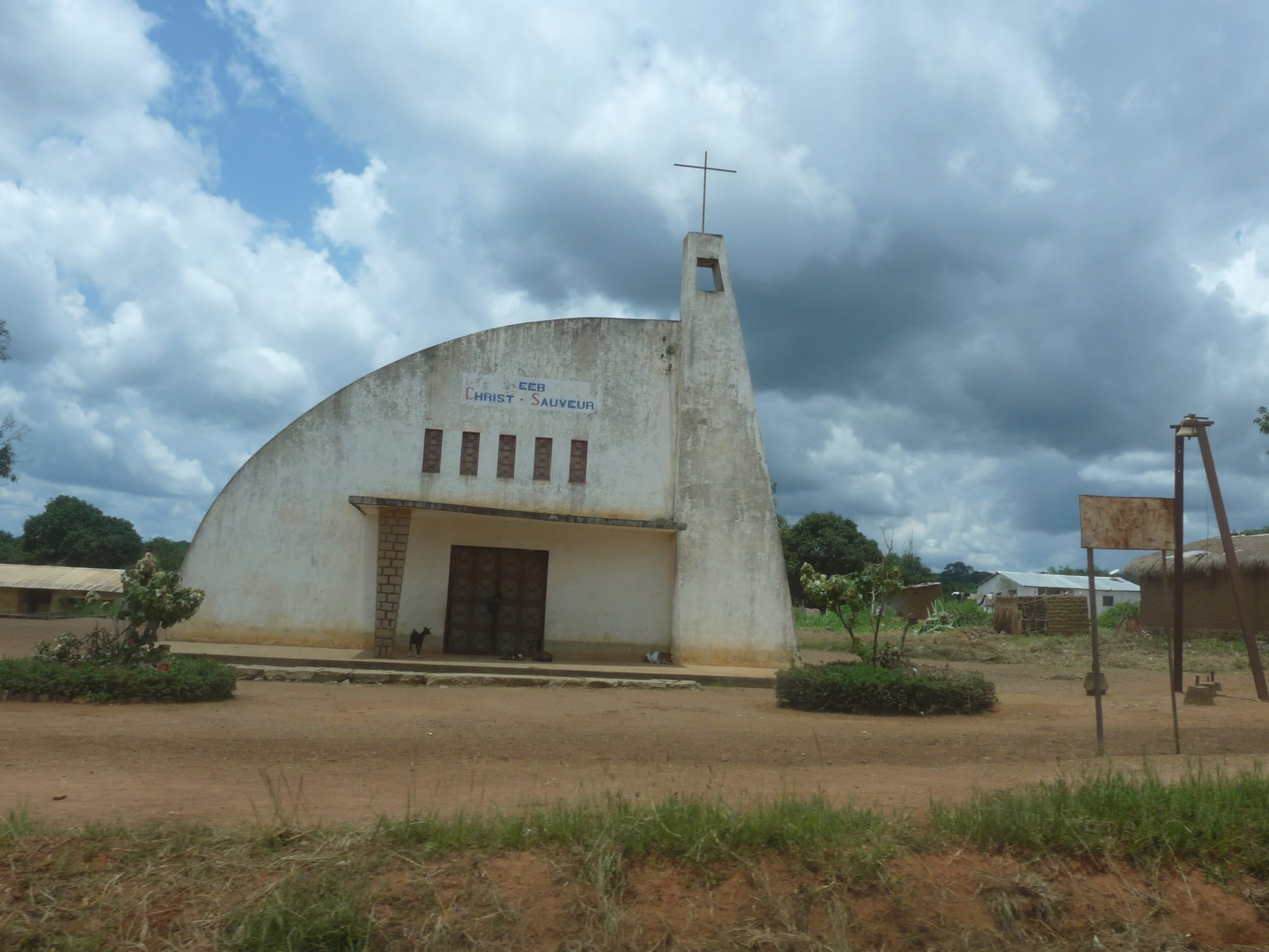 A whitewashed church with a curved gable, a slender bell tower topped by a cross and a row of small windows above a wooden door, set on a red earth square under a cloudy sky
