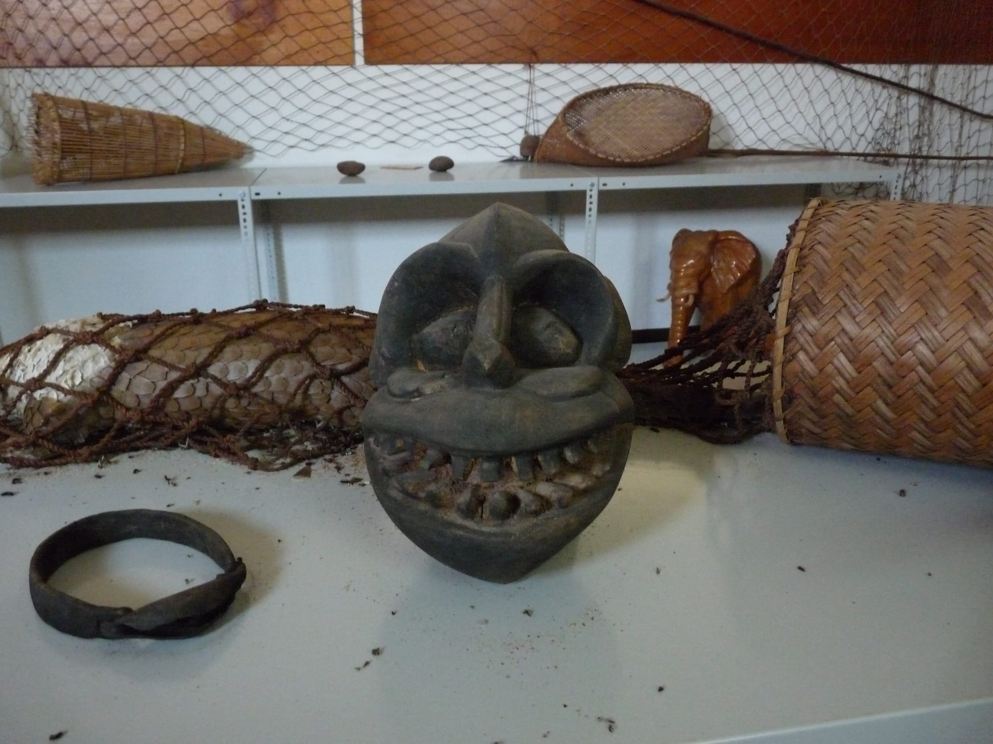 A dark carved wooden mask with large eyes and bared teeth on a table, surrounded by woven baskets, a conical fish trap, a small carved elephant, a netted gourd and a metal bracelet