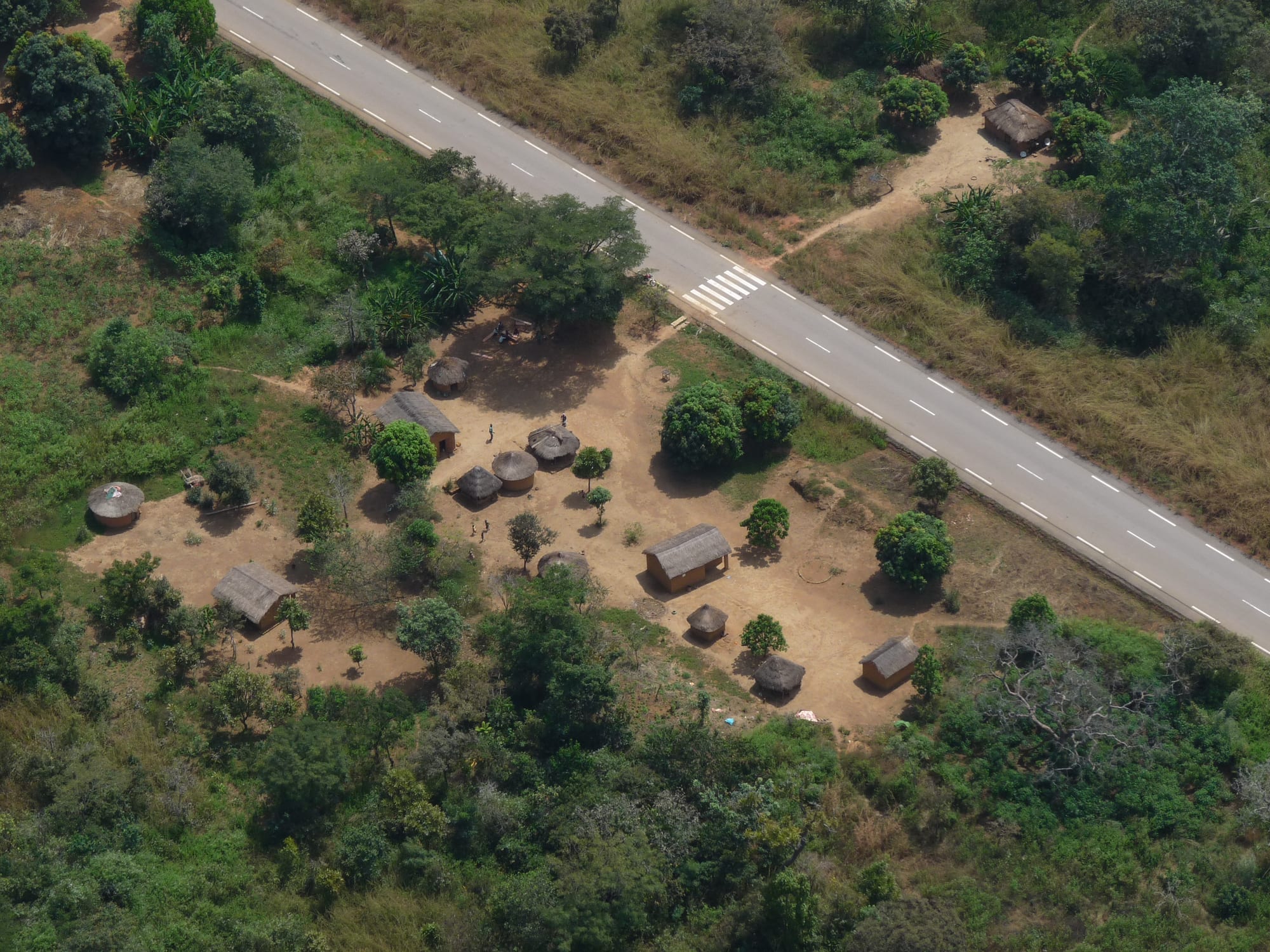 Aerial view of a small cluster of thatched round huts and a few tin-roofed houses on bare red earth beside a paved road, surrounded by dense green bush