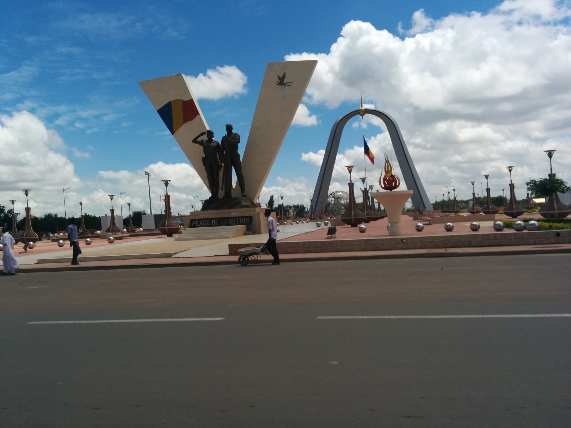 A wide city plaza with a monument and a triumphal arch, traffic passing around it