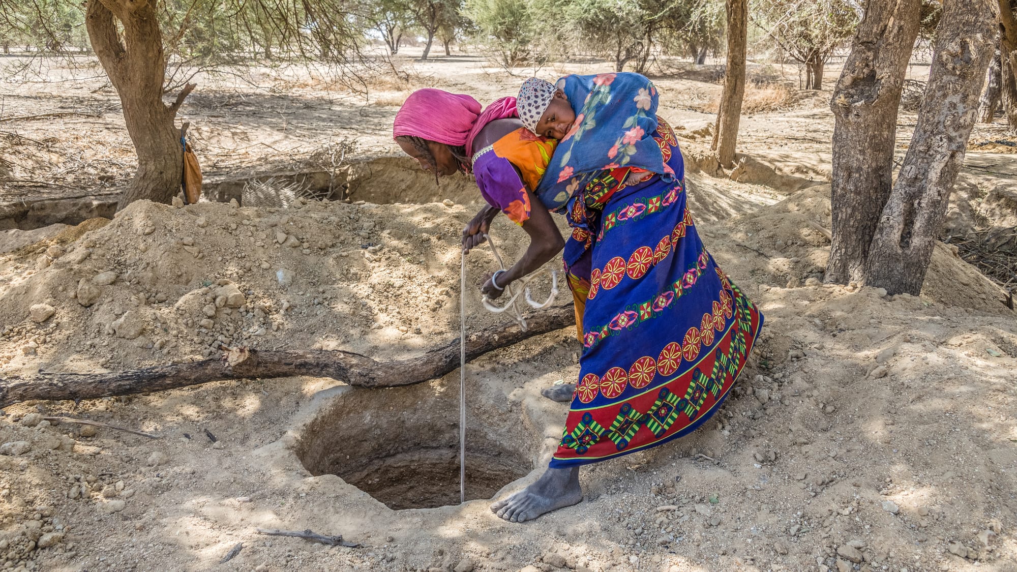 A woman in colourful dress, carrying a child on her back, lowering a rope into a hand-dug well in the Sahel