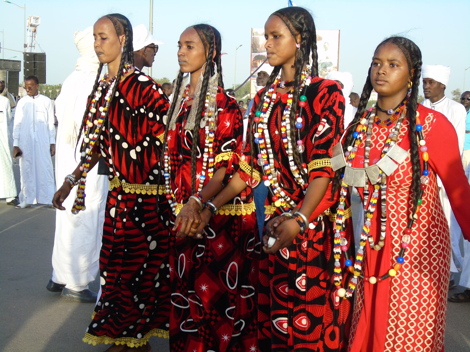 Four young women in red and black patterned dresses, with long braids and layered bead necklaces, walk hand in hand in a line, with men in white robes behind them