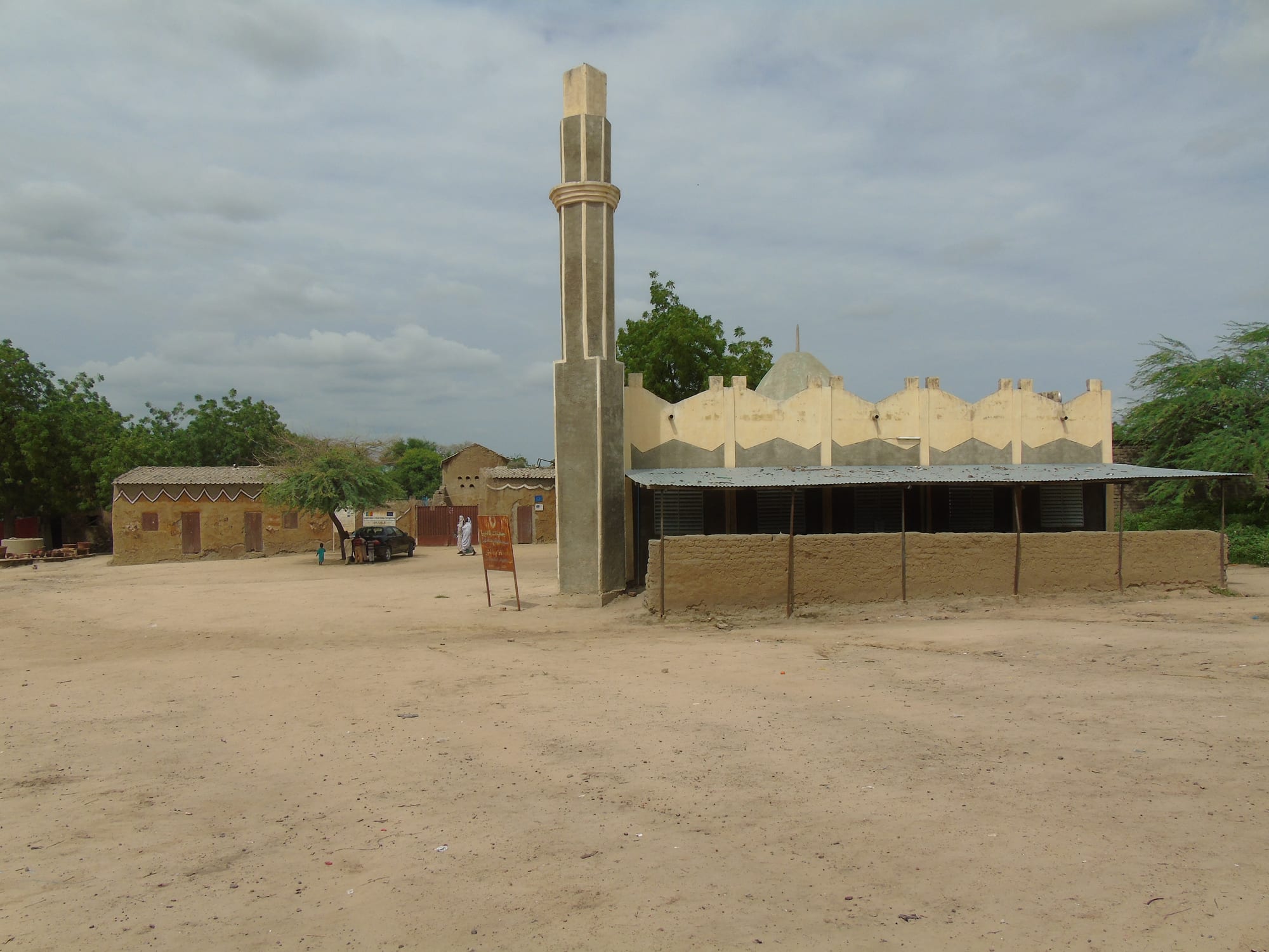 A low mosque with pale scalloped walls and a green-tinged dome beside a tall, square minaret, standing on a wide sandy open space with earth-walled houses behind