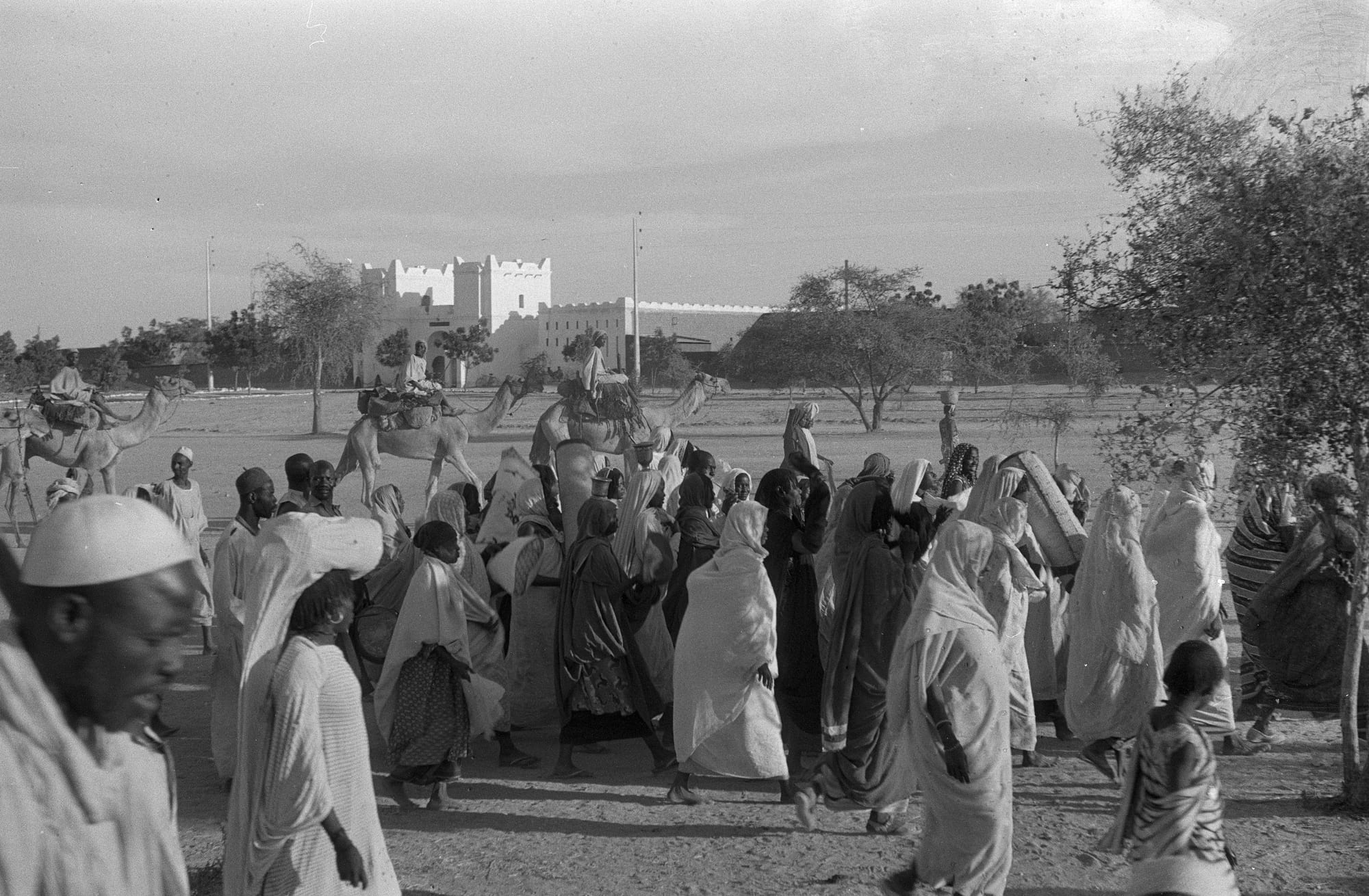 A black-and-white photograph of a crowd of women in long white wraps walking across a sandy plain, with three men riding camels behind them and a white building with battlements in the distance