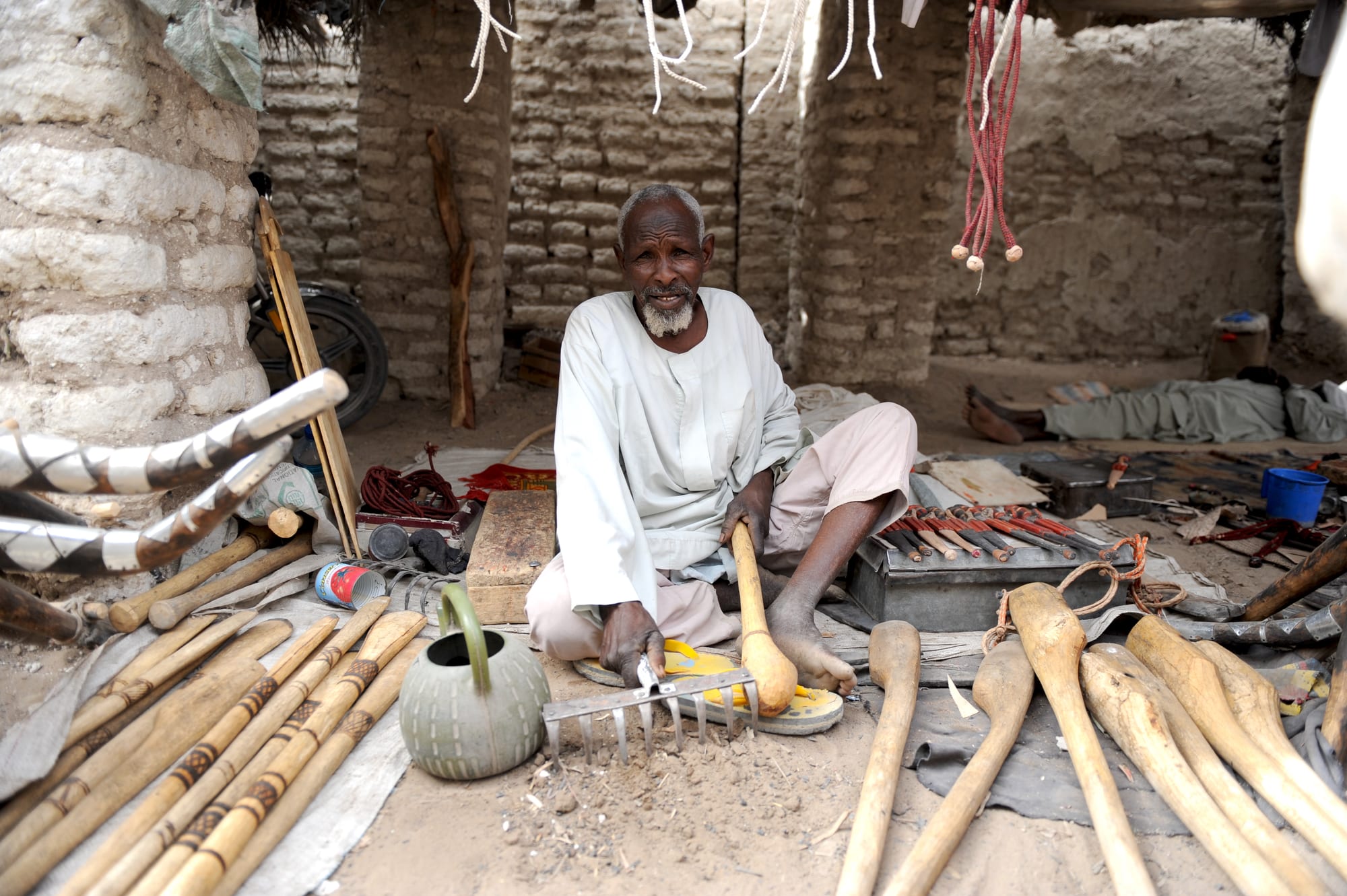 An older man in a white robe sits cross-legged in a mud-brick workshop, surrounded by carved wooden handles, tools and a green metal kettle