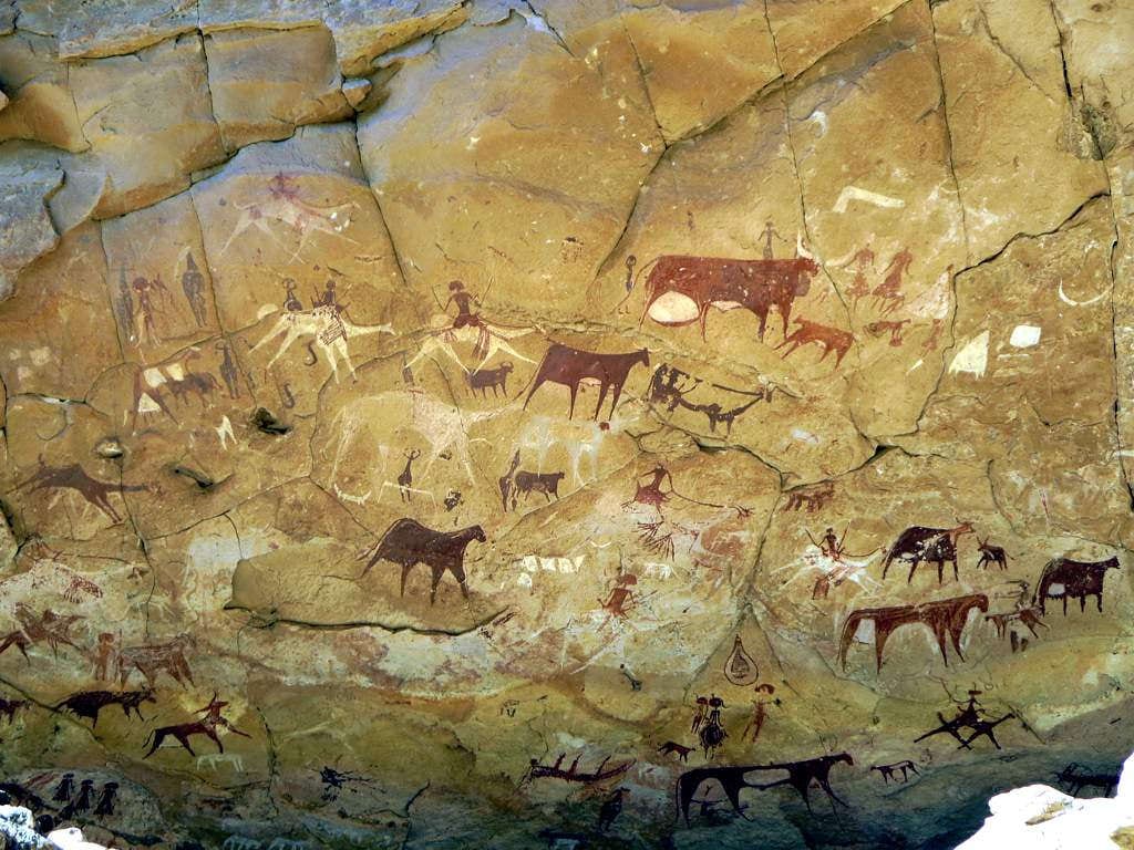 Faded ochre paintings of cattle and human figures on a pale sandstone wall in a desert cave