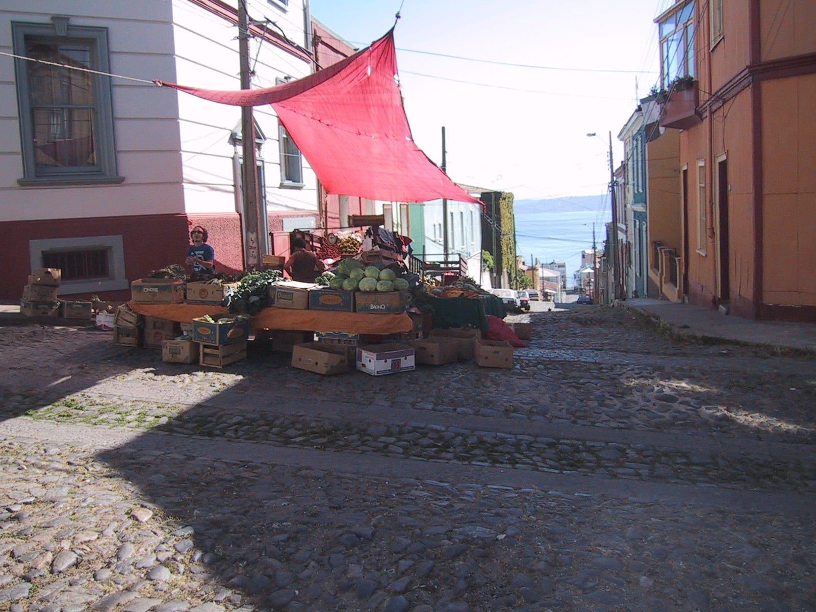 A small fruit and vegetable stall shaded by a red awning on a steep cobblestone street in Valparaíso, with the sea visible below