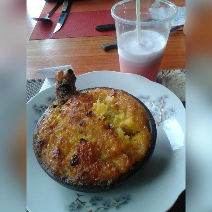 A baked corn casserole in a clay dish, beside a glass of a pale, frothy drink