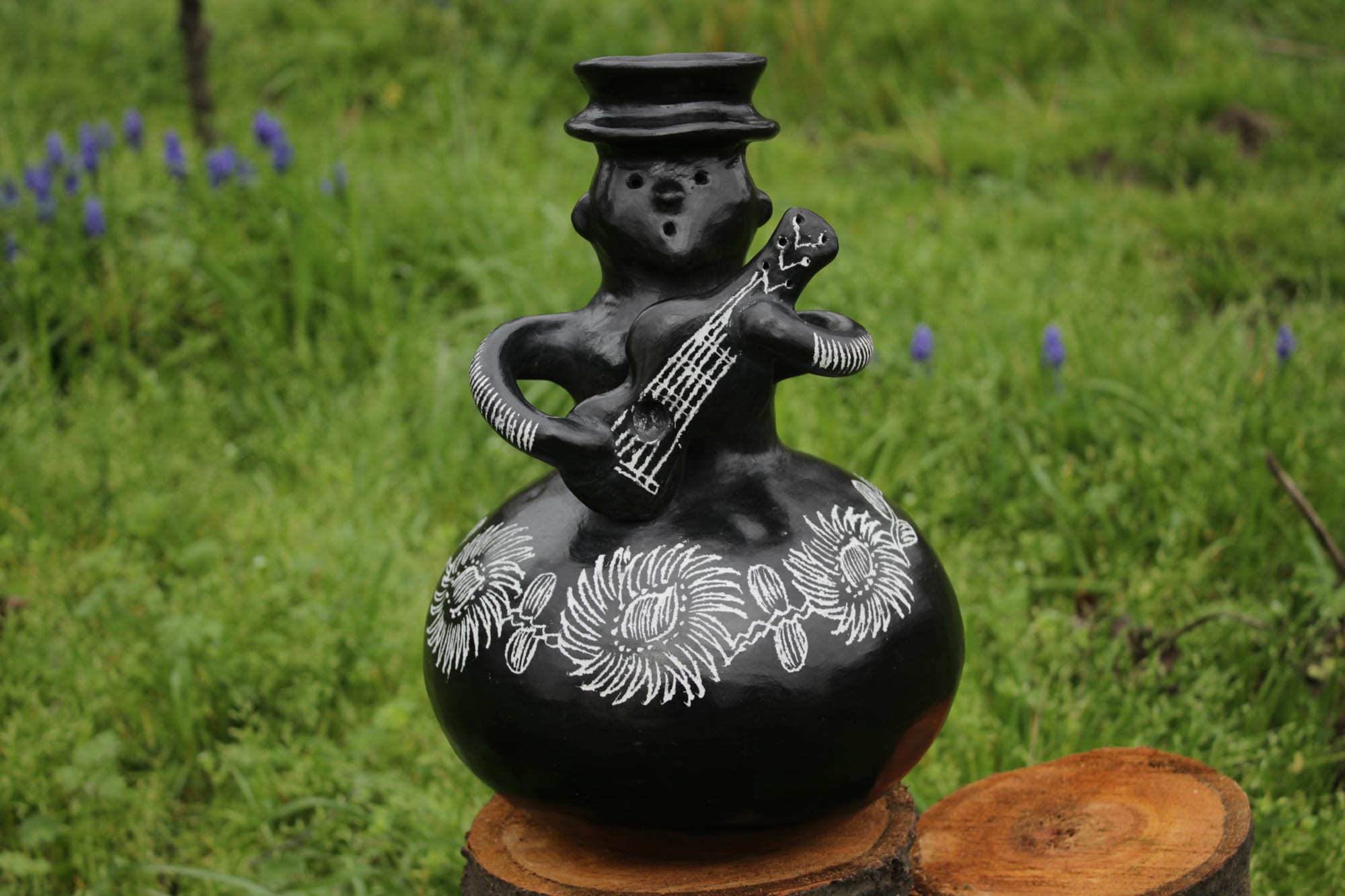 A glossy black clay figure of a woman in a hat playing a guitar, seated on a round jug decorated with white incised flowers, displayed on a tree stump against green grass