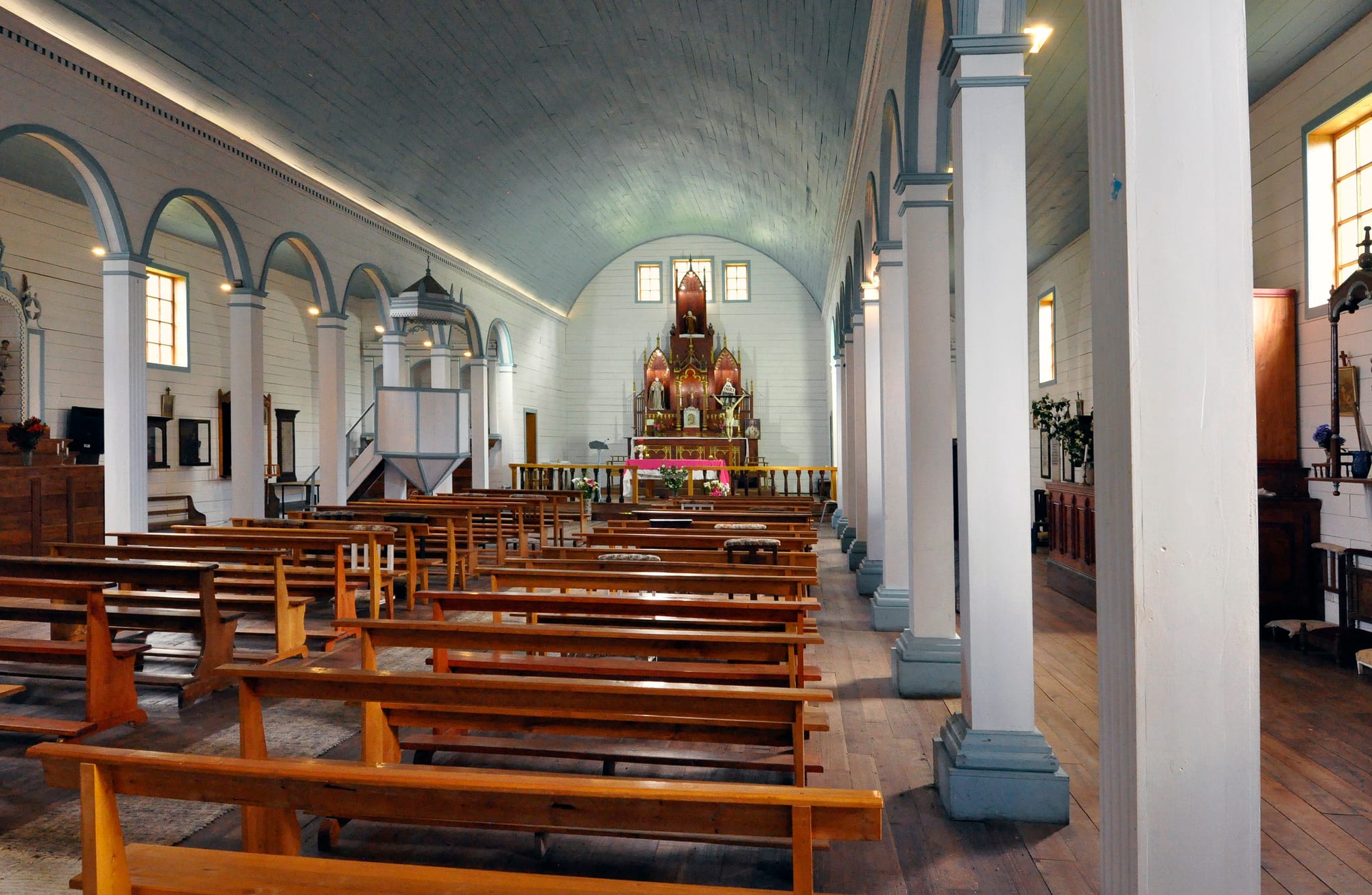 The interior of a wooden church with rows of wooden pews, white painted columns and arches, a curved blue-grey wooden ceiling and a carved altar at the far end