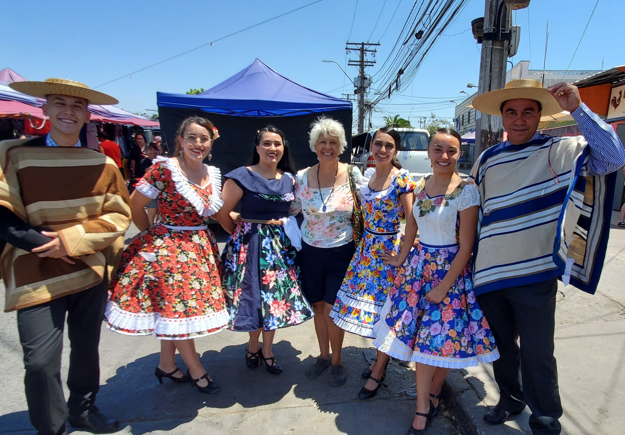 Two men in wide-brimmed straw hats and striped ponchos stand at either end of a row of smiling women in full, flowered cueca dresses, on a sunny street lined with market stalls