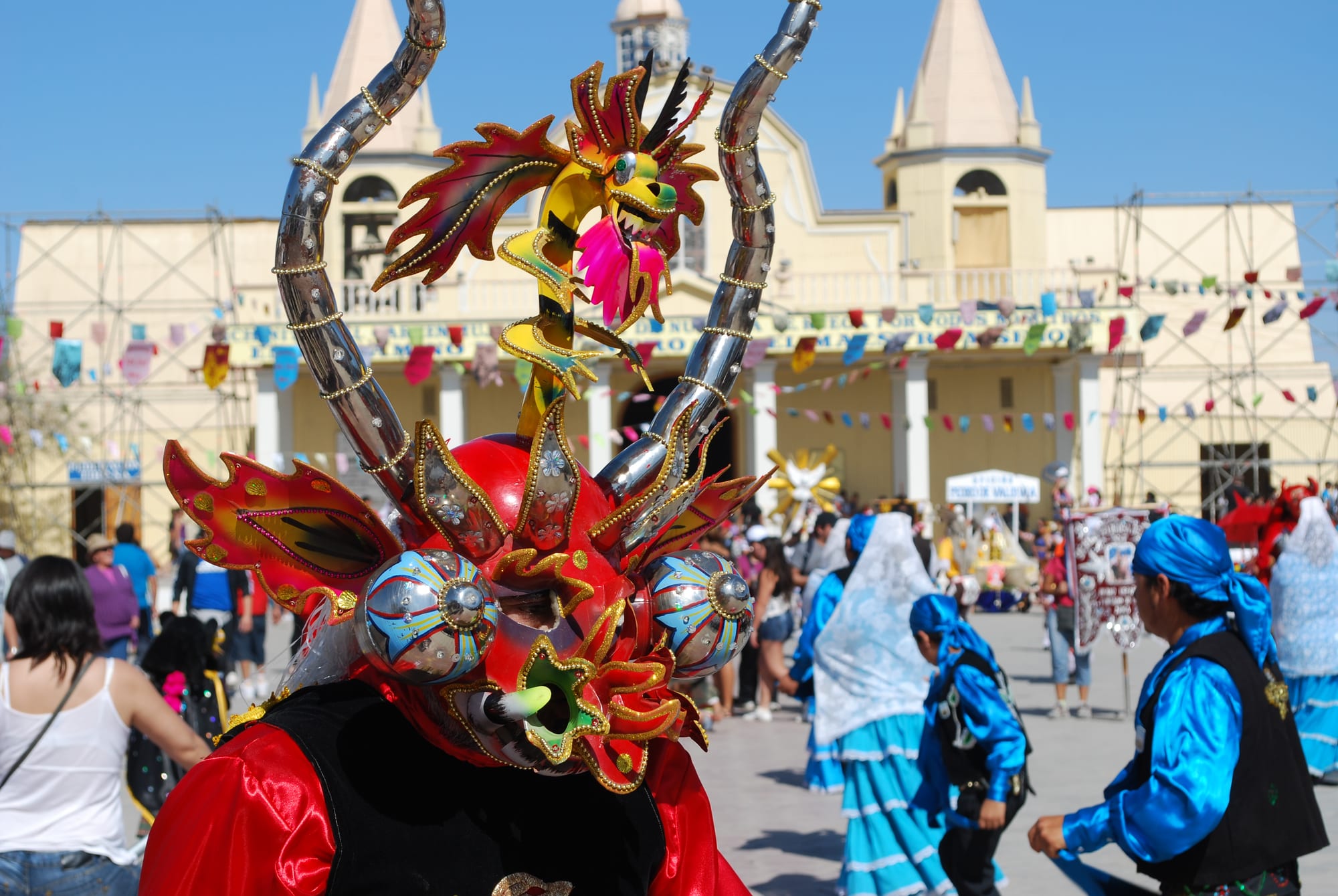 A dancer in a large red devil mask with silver horns, a small dragon and glittering decoration stands in front of a pale church, with dancers in bright blue and white costumes behind