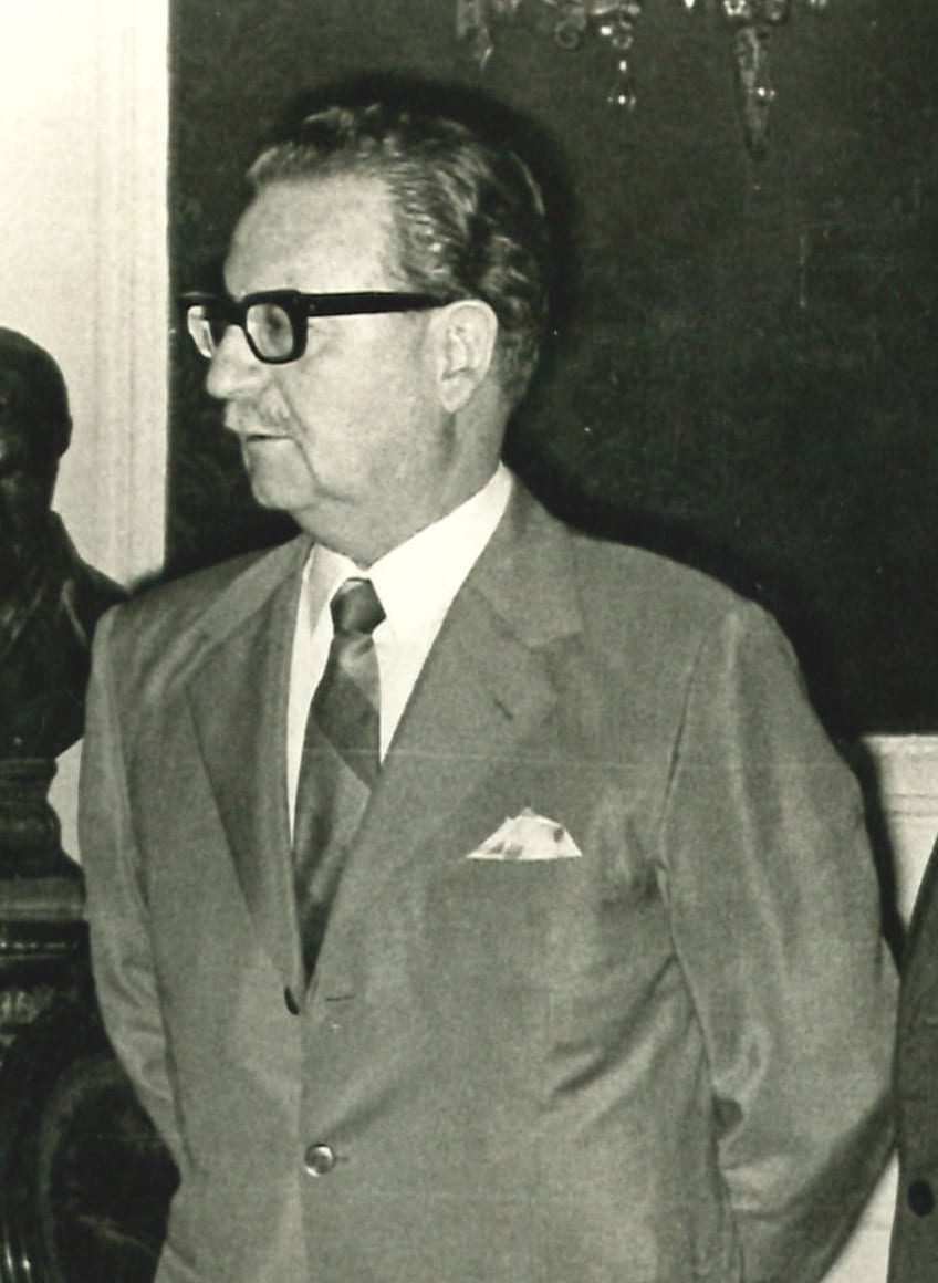 Salvador Allende in glasses and a suit, seated at a desk, looking to one side
