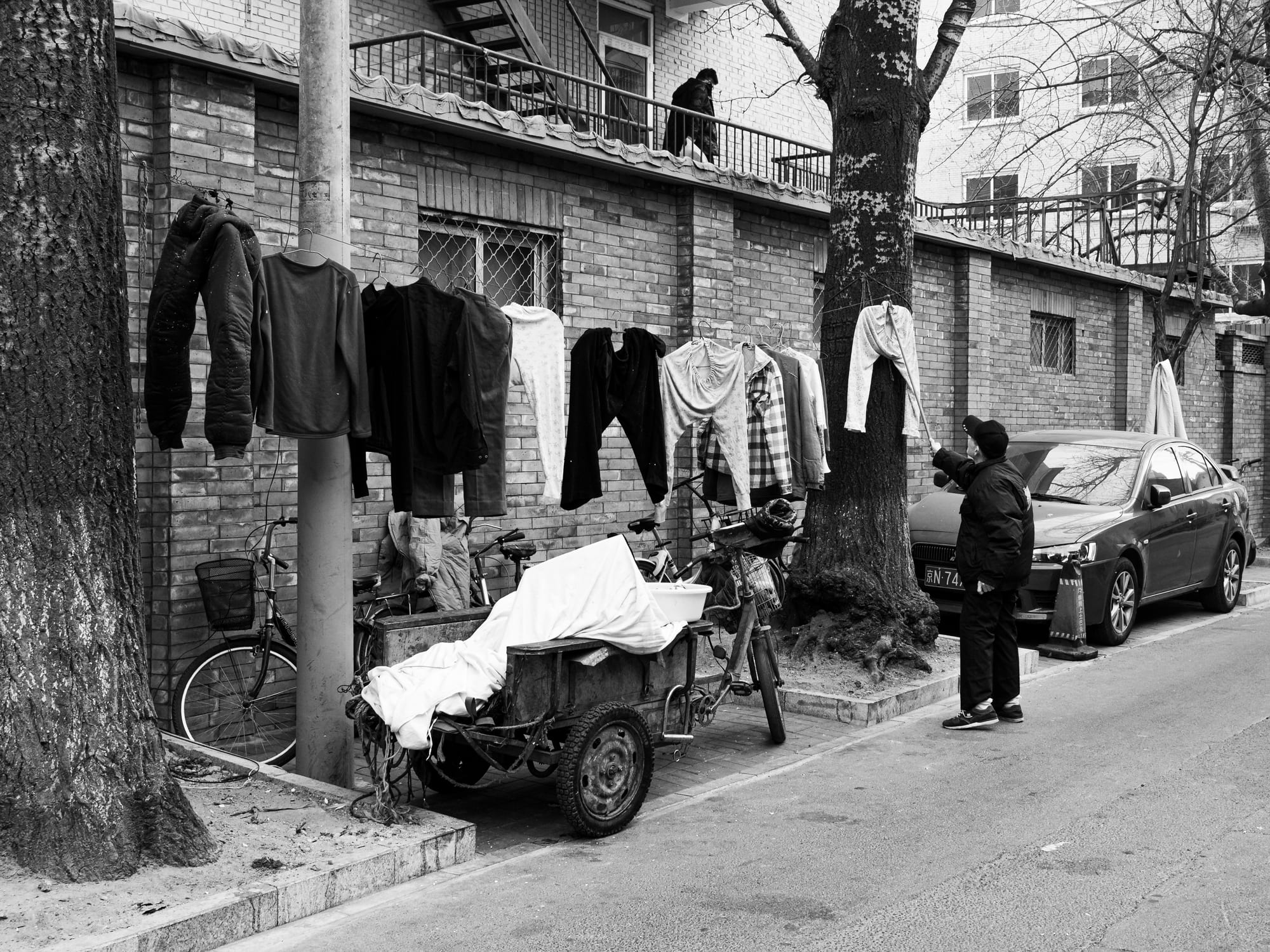 A black and white street scene of a Beijing hutong alley, with laundry hung on lines between trees and an elderly resident tending to it