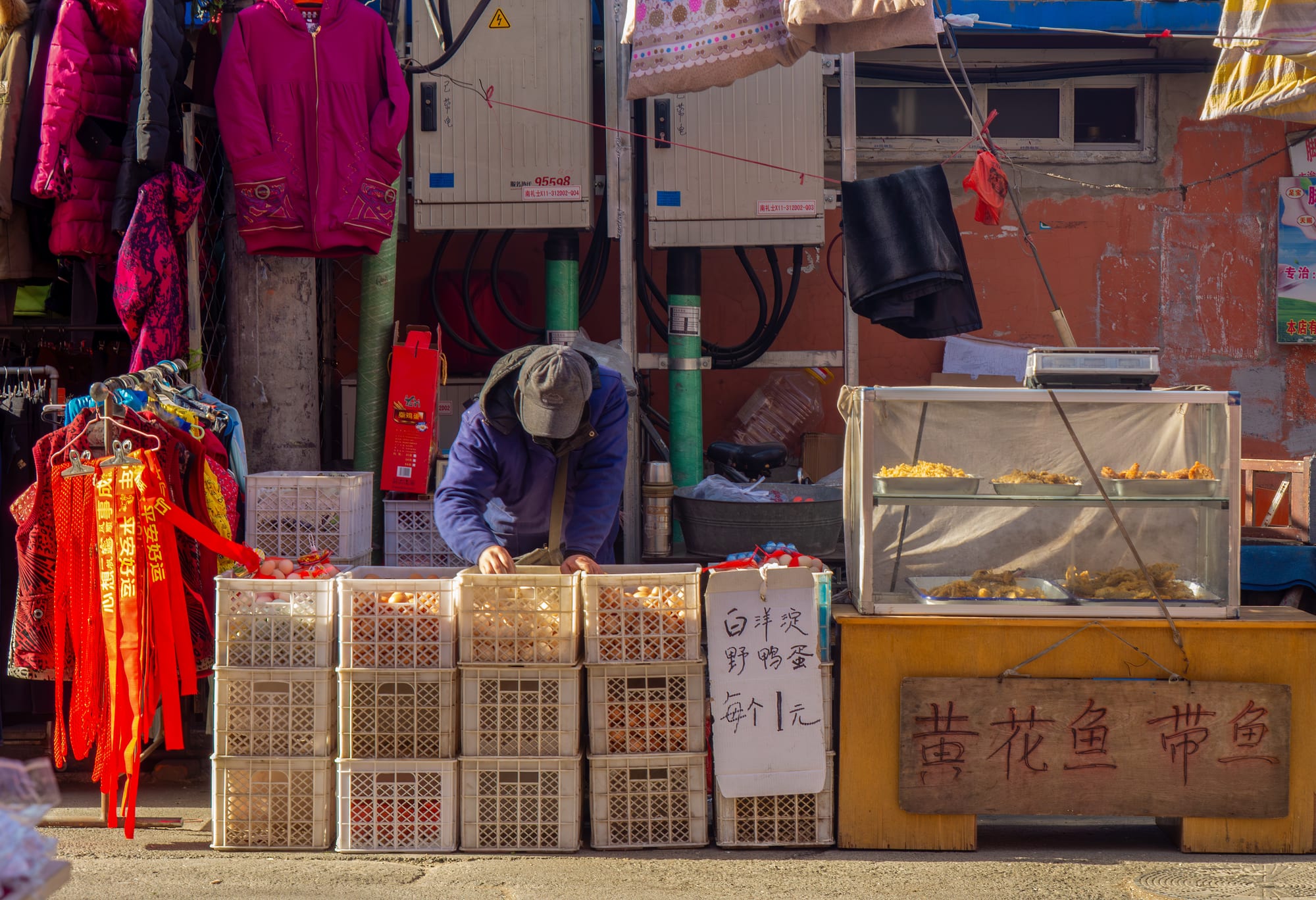 A street vendor arranging crates of eggs at an outdoor market stall in Beijing, with clothing and food stalls around