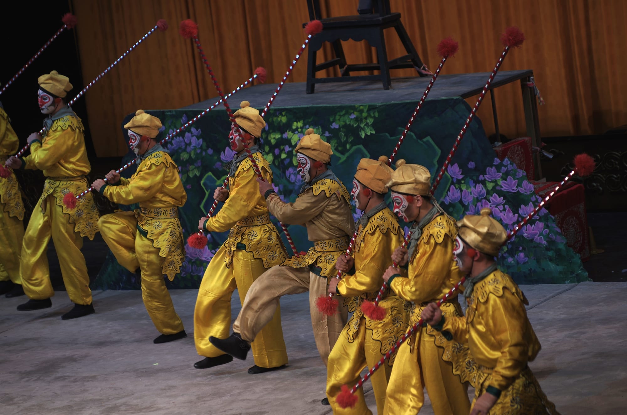 A row of performers in bright yellow costumes and caps, with white and red painted monkey faces, holding long red and white striped staffs on a stage