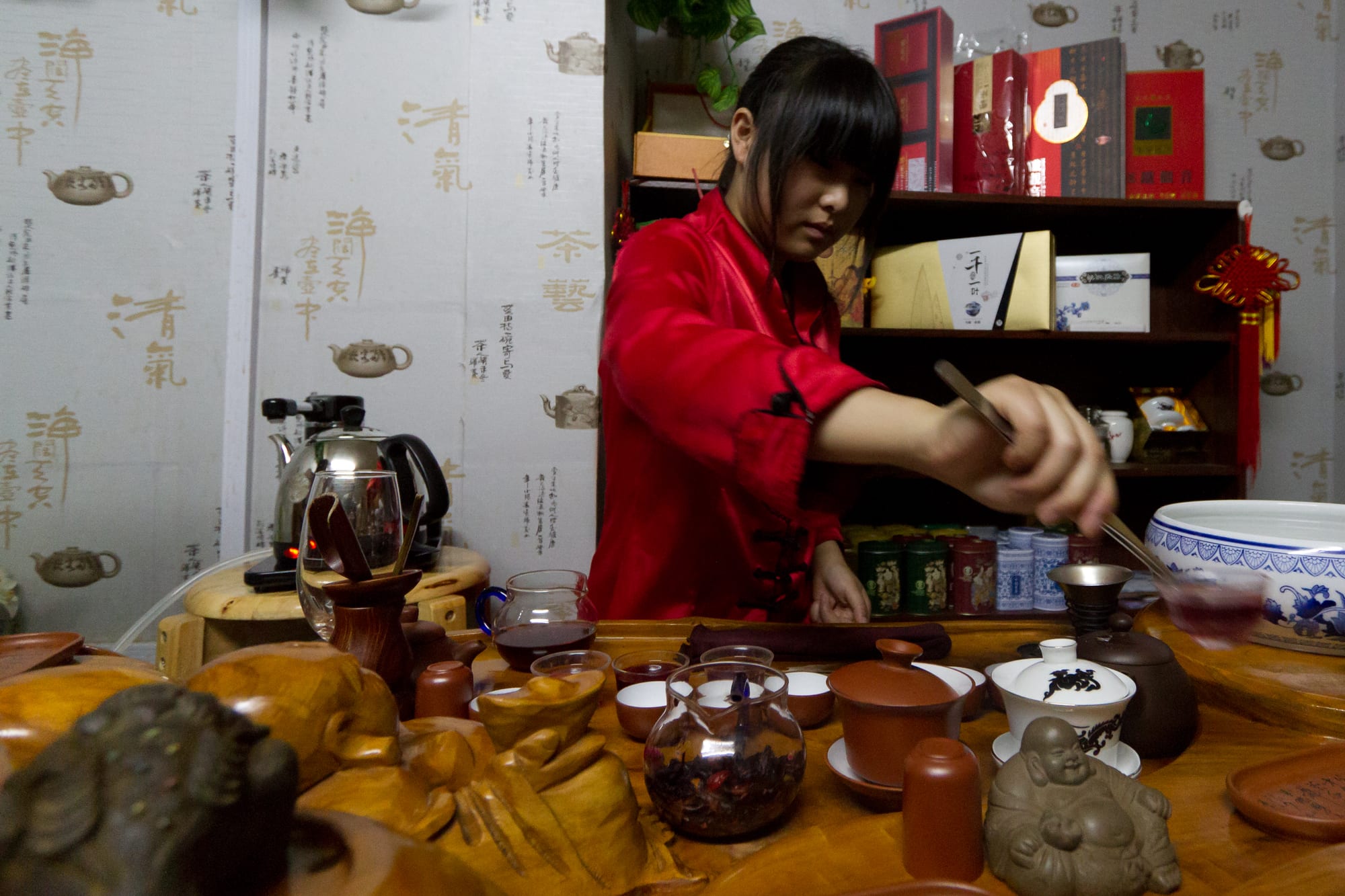 A woman in a red jacket uses tongs to lift a small cup at a carved wooden tea table crowded with clay teapots, glass pitchers and tiny cups, with boxes of tea on shelves behind her