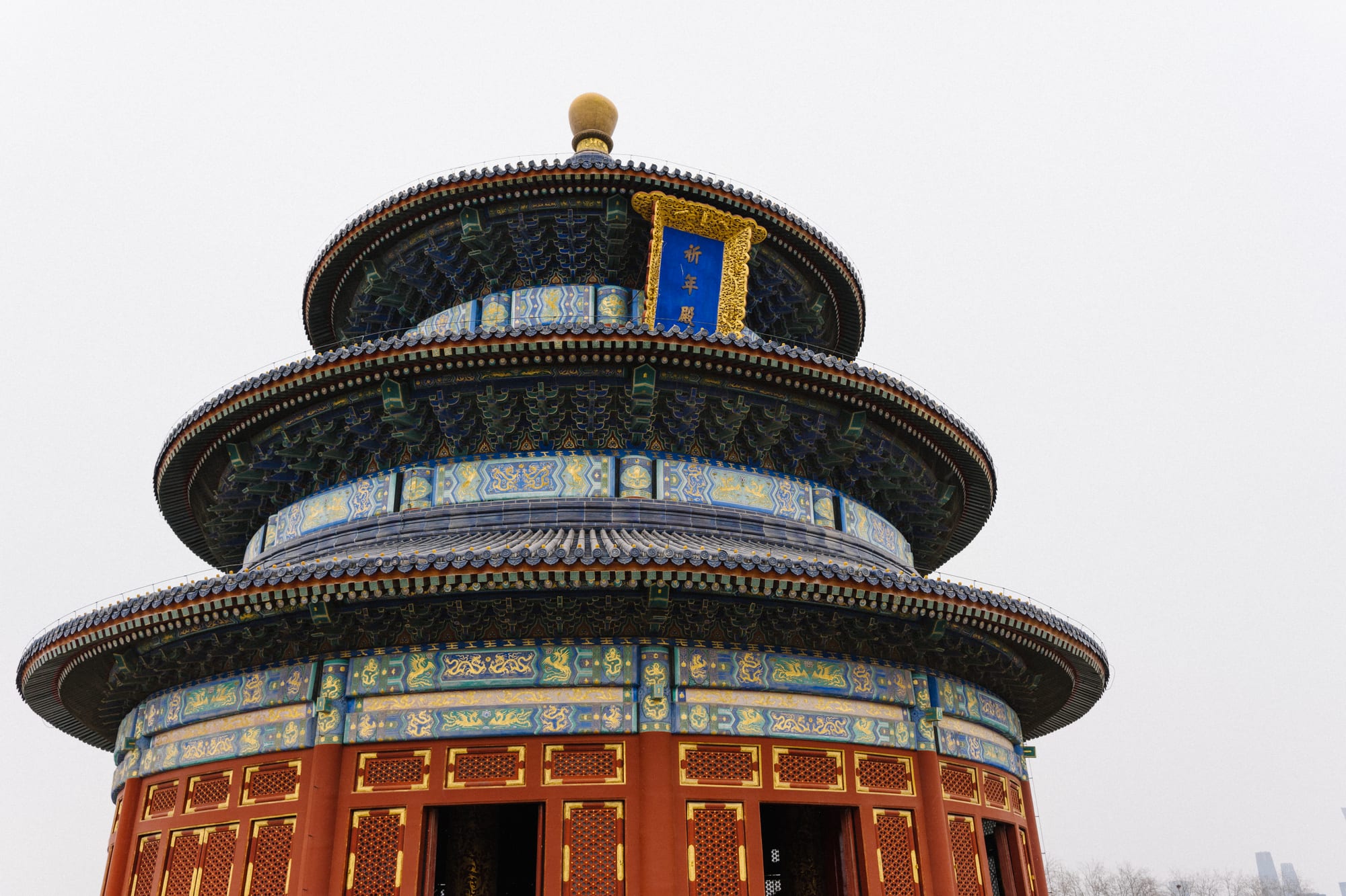 A round three-tiered wooden hall with dark blue tiled roofs, painted blue, green and gold eaves, a gilded finial and red lattice doors, under a pale winter sky