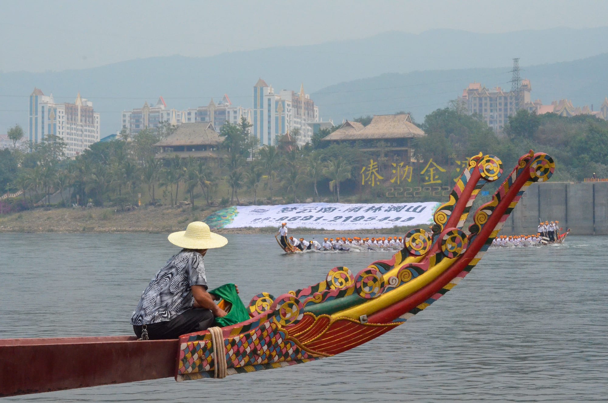 The carved and brightly painted prow of a dragon boat in the foreground, with a woman in a straw hat sitting on it, and long boats of rowers in white racing on the river beyond
