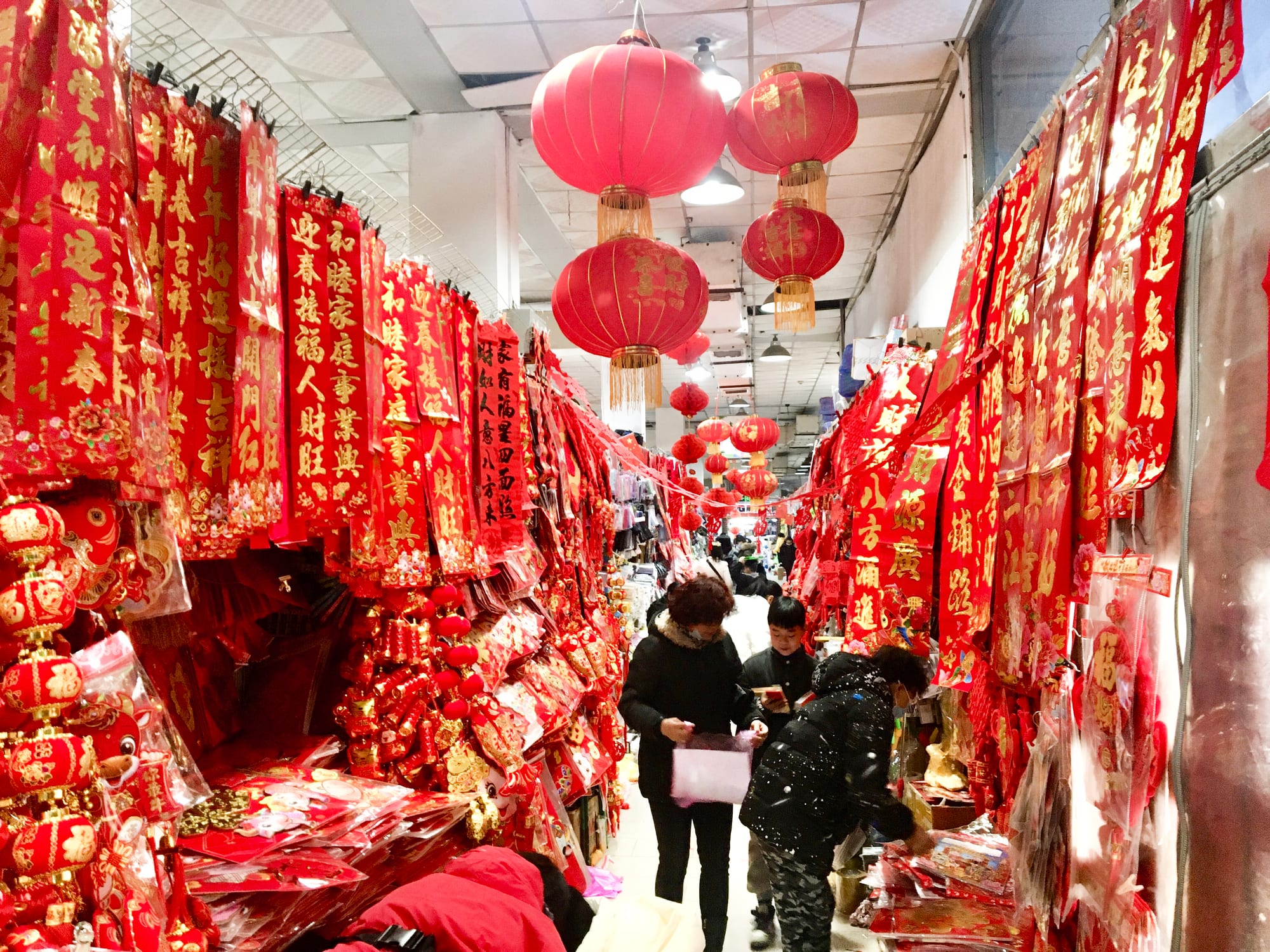 A market aisle hung from floor to ceiling with red and gold paper couplets and decorations, with large red lanterns overhead and shoppers walking between the stalls