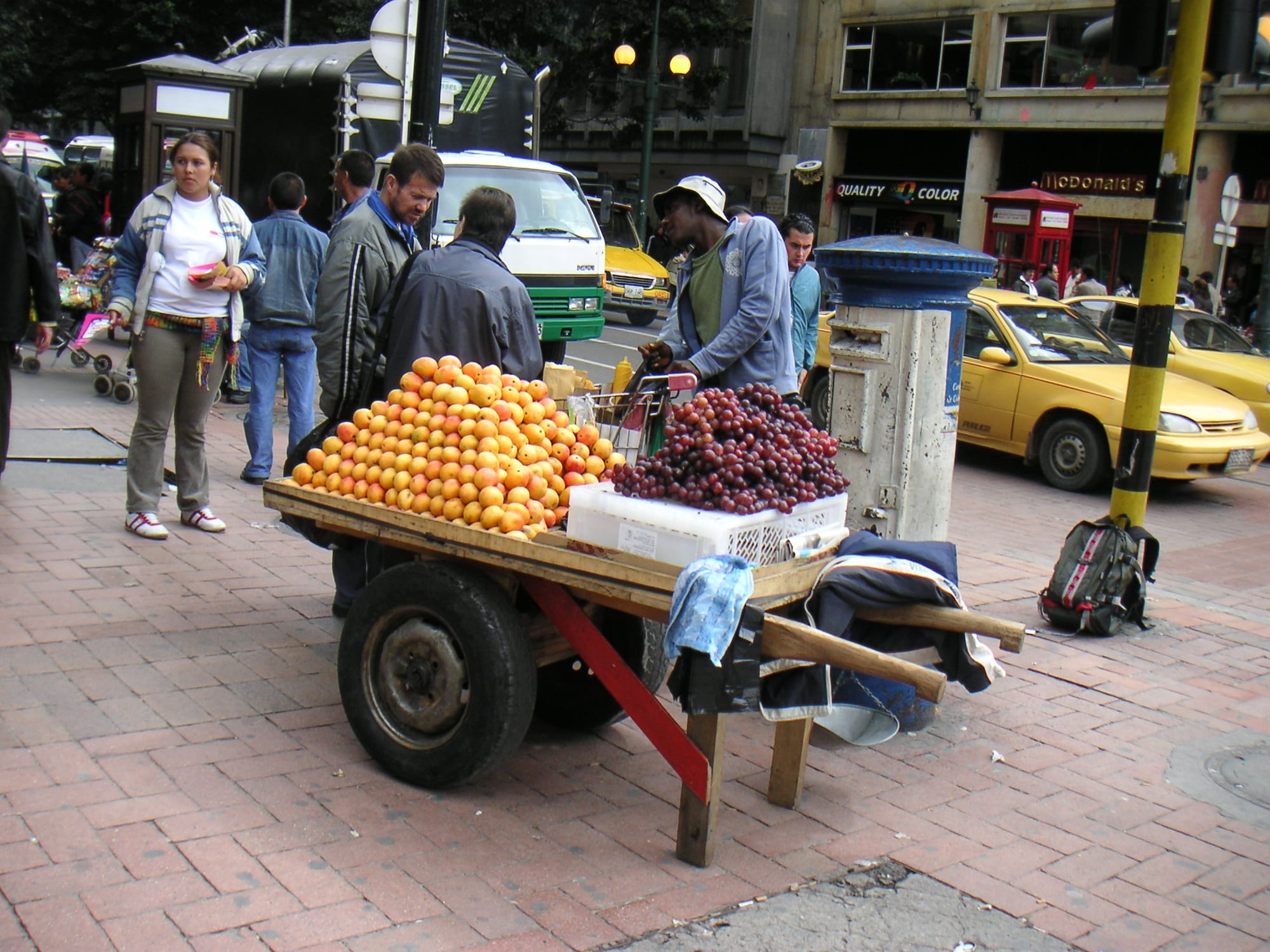A street fruit vendor's cart piled high with oranges and grapes on a city pavement, with pedestrians and a yellow taxi behind