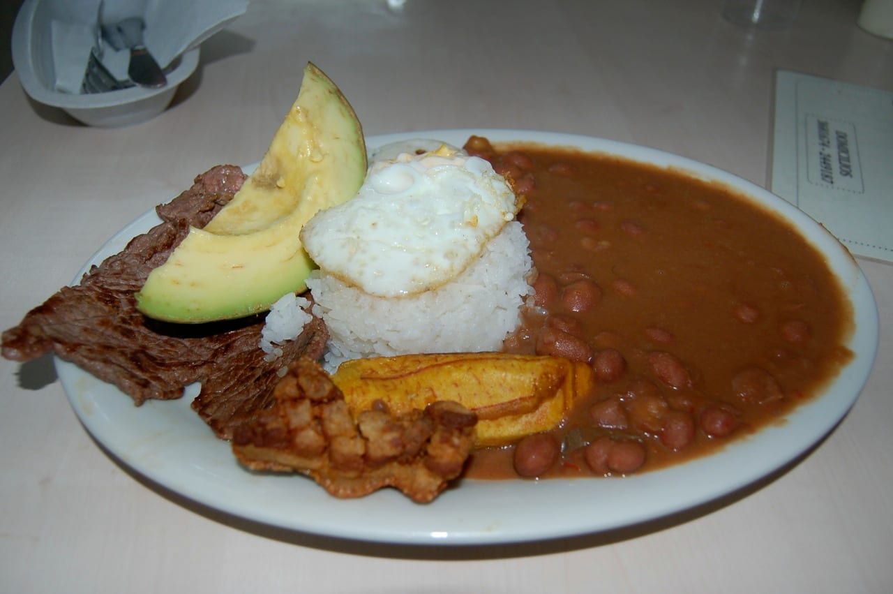 A large plate loaded with rice, beans, ground beef, sausage, fried egg, plantain, avocado and a piece of pork crackling