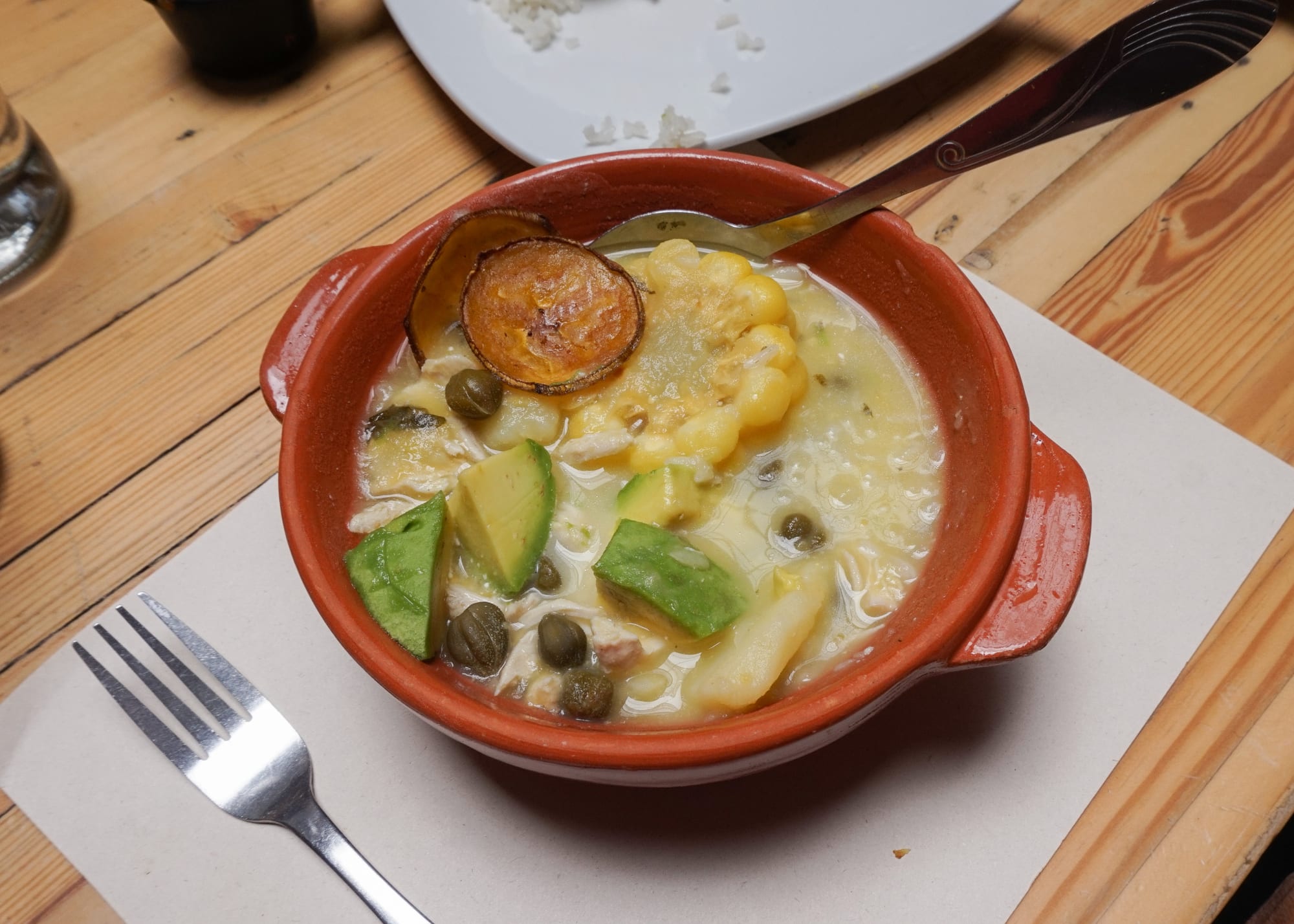 A bowl of thick yellow soup with chicken, corn and potato, served with a side of cream, capers and avocado