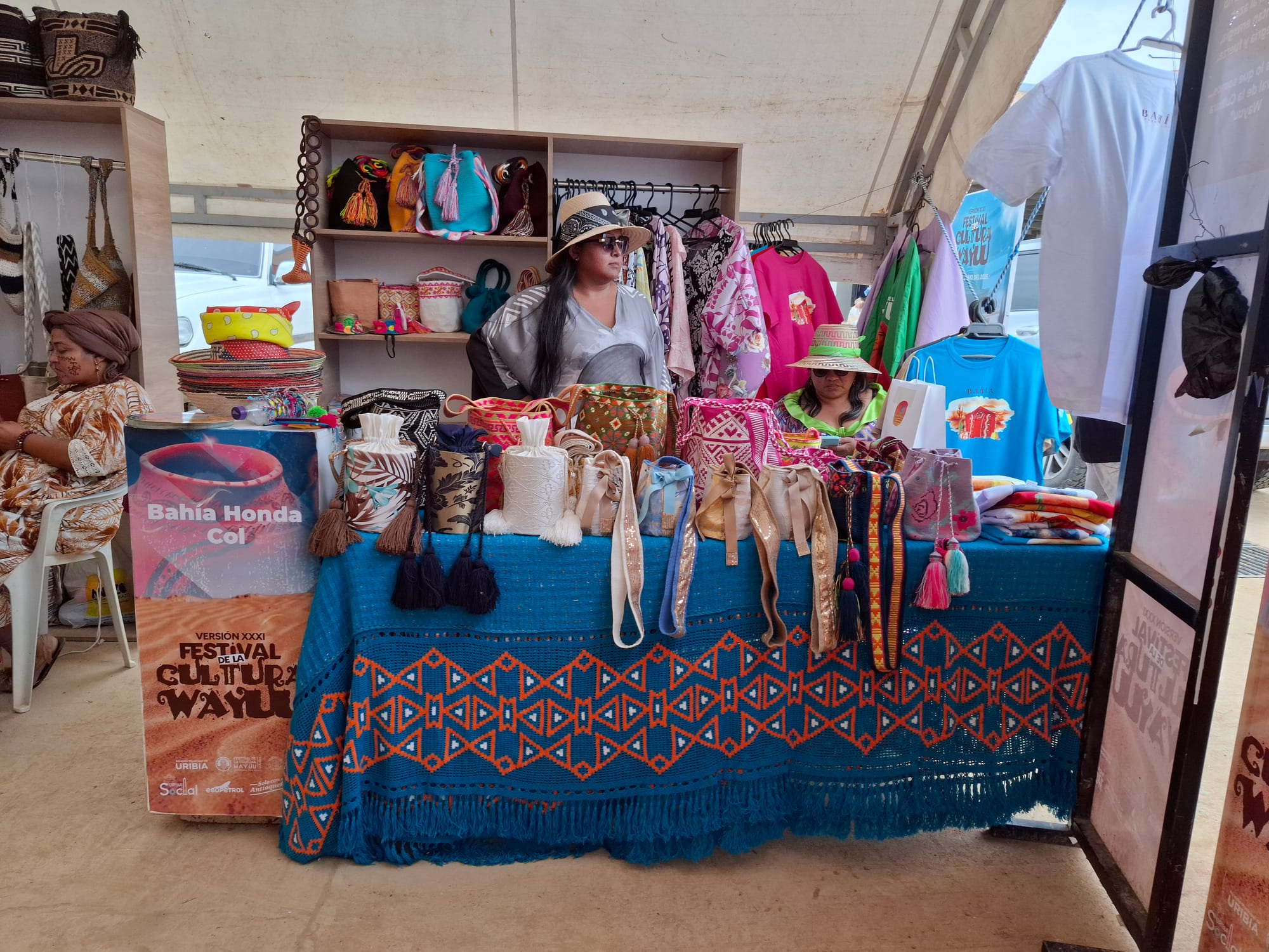 A stall with a table covered in a blue and orange woven cloth, displaying colourful woven bags with tassels, with more bags and hats on shelves behind two women in hats