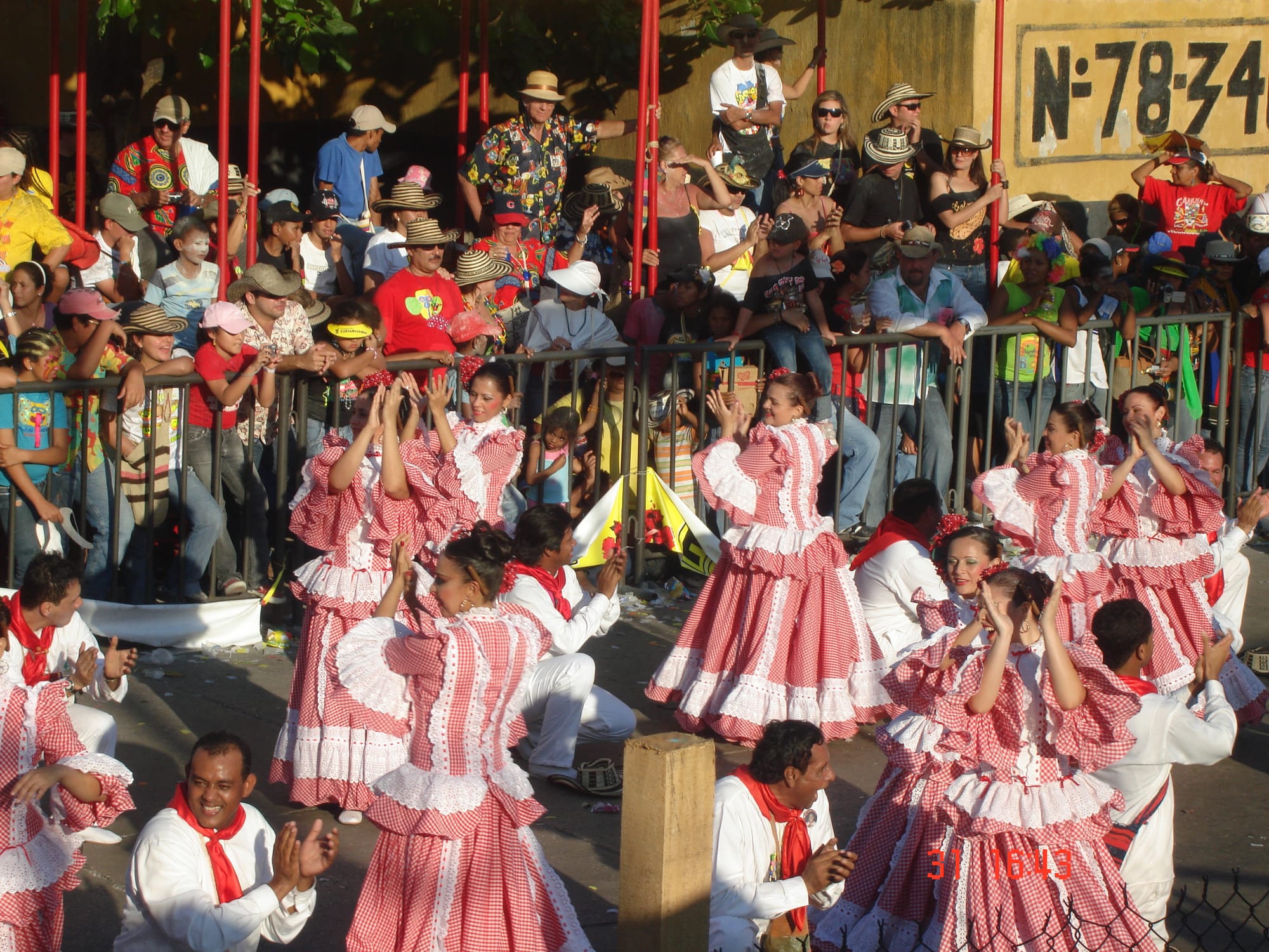 Women in full red and white checked dresses dance with raised arms while men in white shirts and red neckerchiefs clap, in front of a crowd of spectators in straw hats behind a barrier