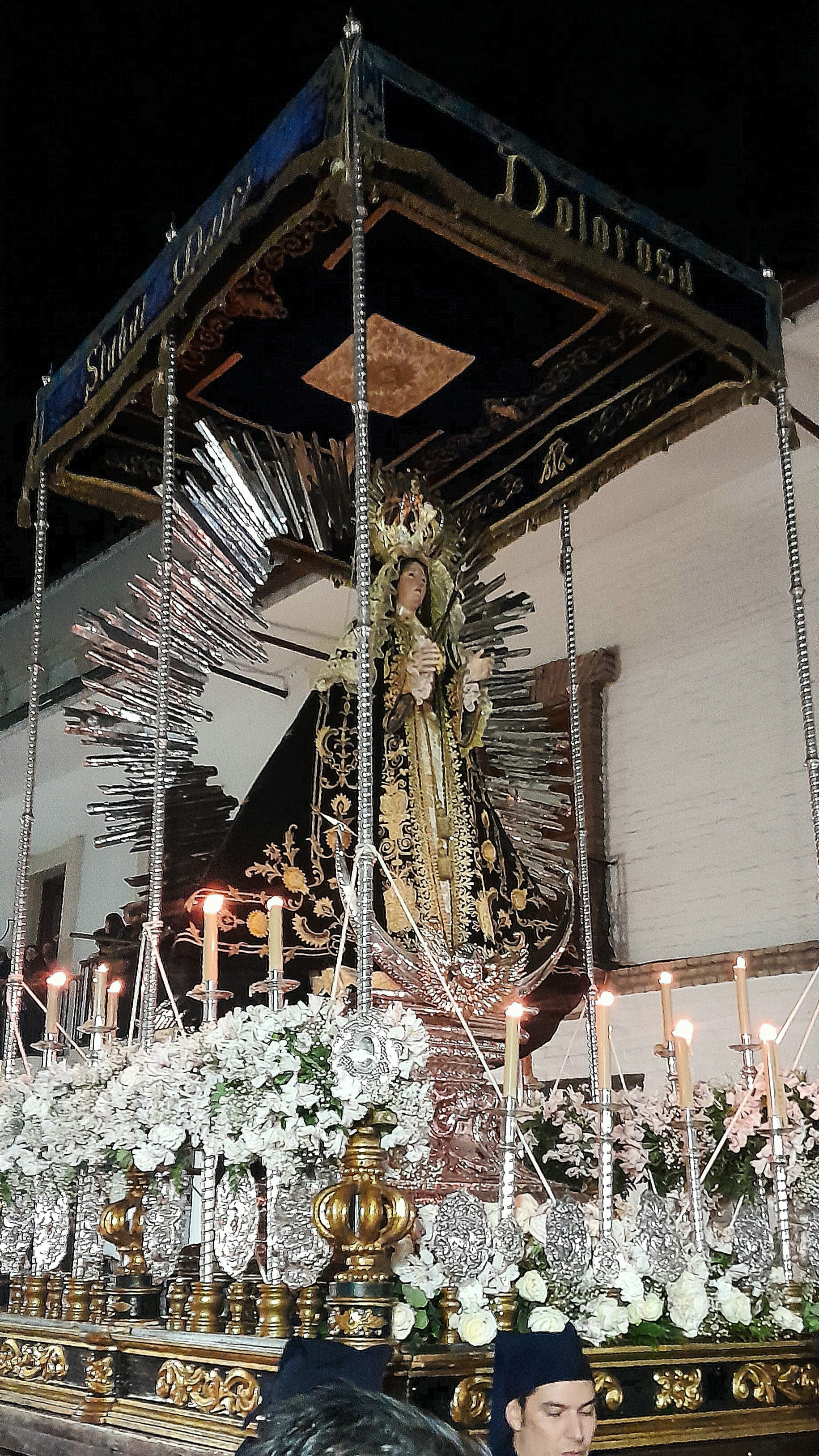 A carved statue of the Virgin Mary in a black and gold embroidered robe, framed by silver rays and a dark canopy, on a float decked with white flowers and lit candles, carried through a street at night