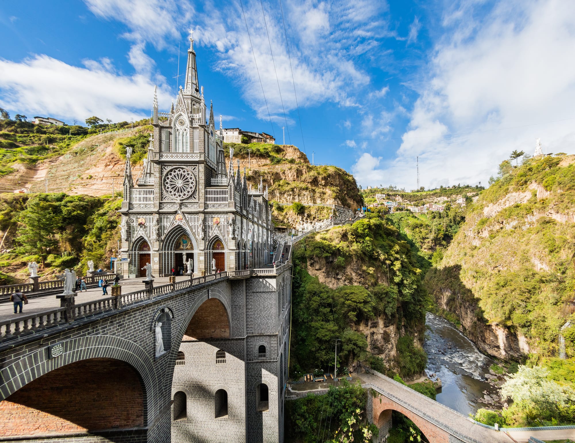 A grey stone neo-Gothic church with a tall spire and a large rose window, built on an arched bridge across a deep green river gorge under a blue sky