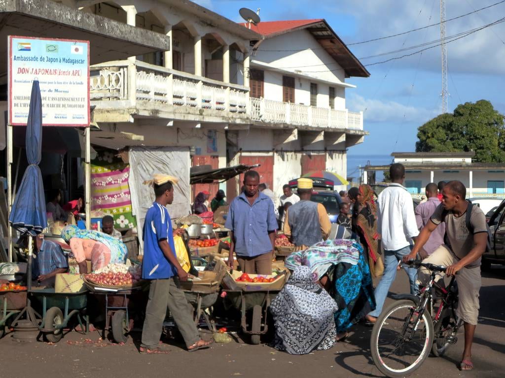 A busy street market in Moroni with vendors, produce laid out on carts, a cyclist, and a colonial-era building behind