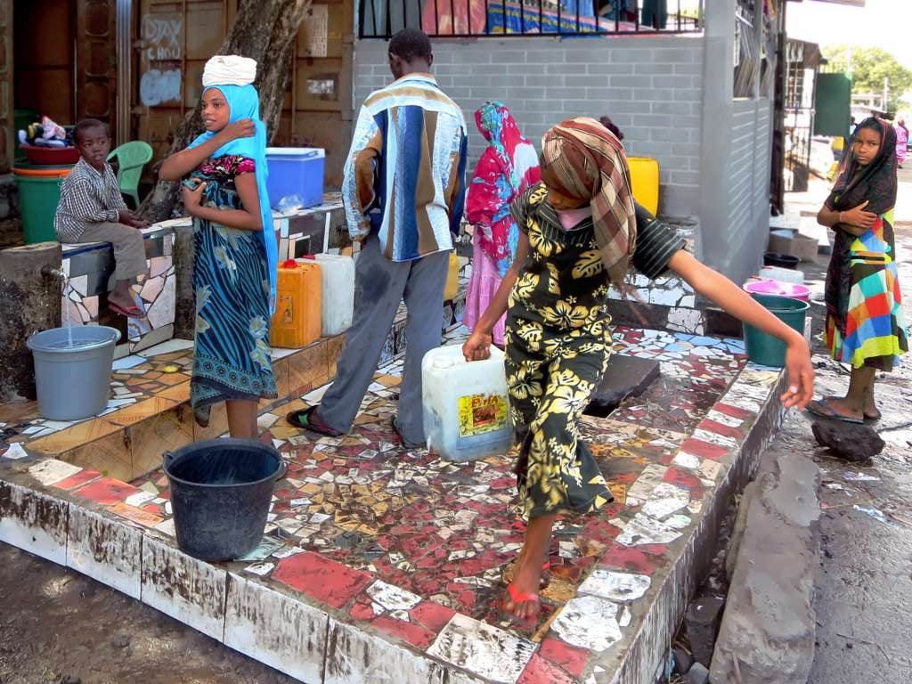 Children and adults filling buckets and containers at a public water tap in a Moroni street