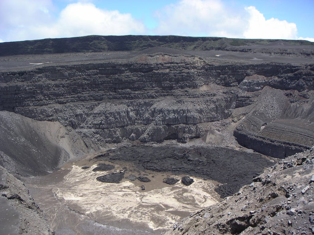 A broad dark volcanic mountain rising from tropical forest under a cloudy sky