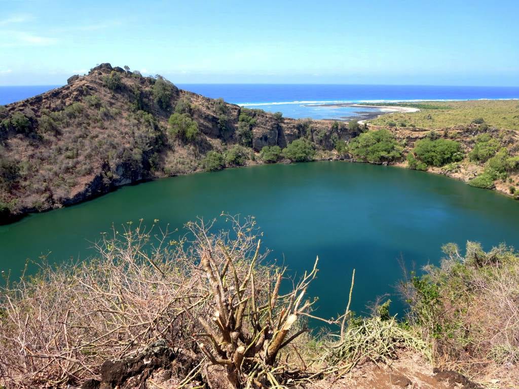 A round, deep crater holding a green-blue lake, ringed by dark vegetated slopes near the sea